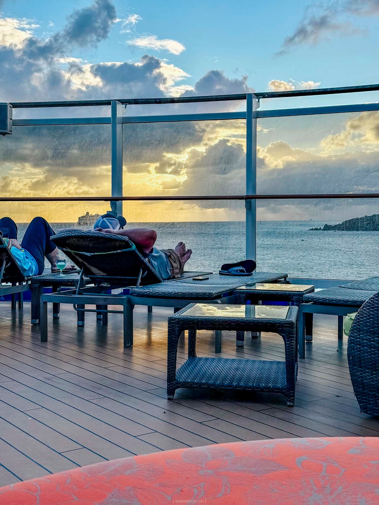 Tourists relaxing on lounge chairs on a cruise ship deck at sunset with ocean views and dramatic clouds.