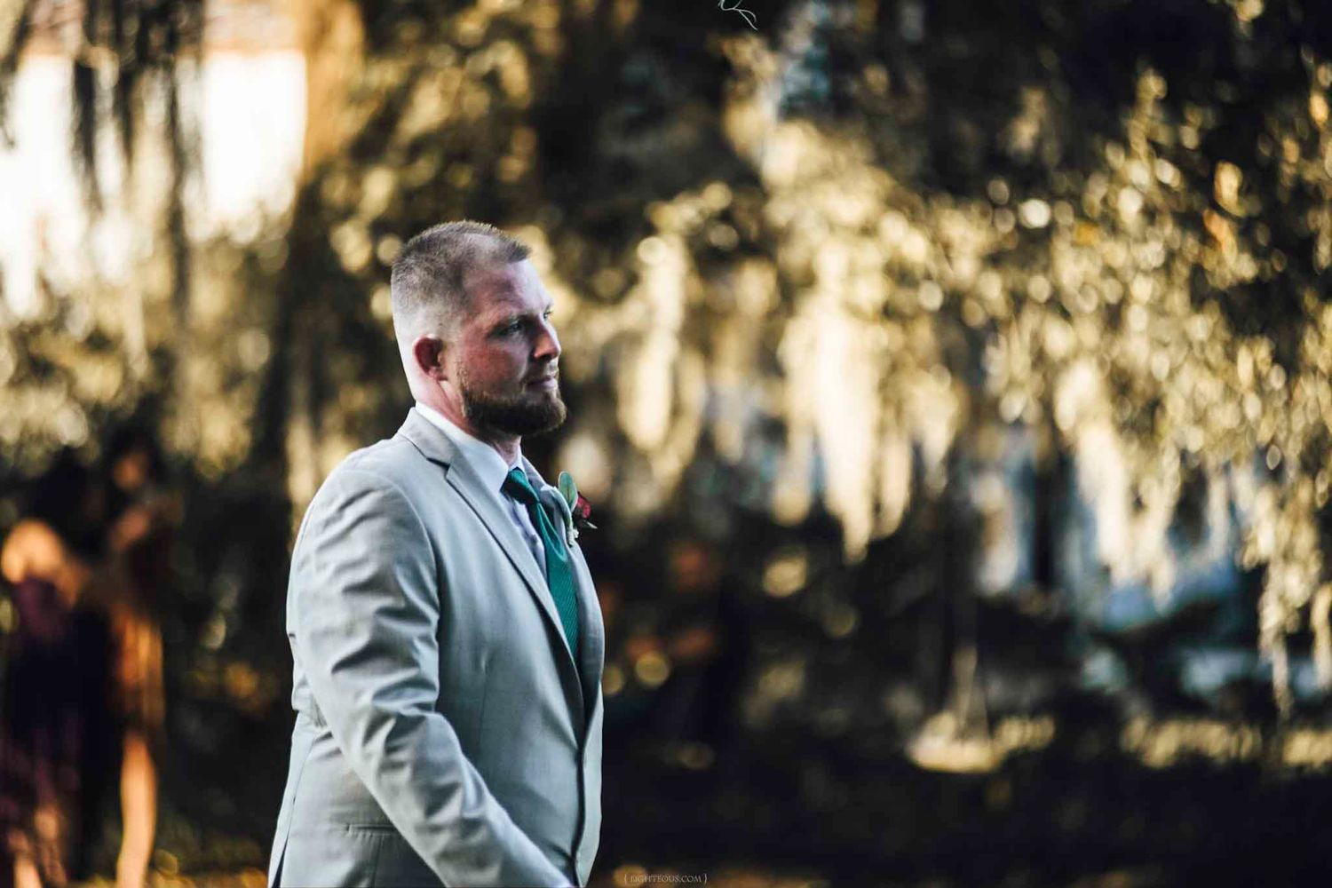Groom in gray suit with green tie standing outdoors among sunlit trees at a wedding.