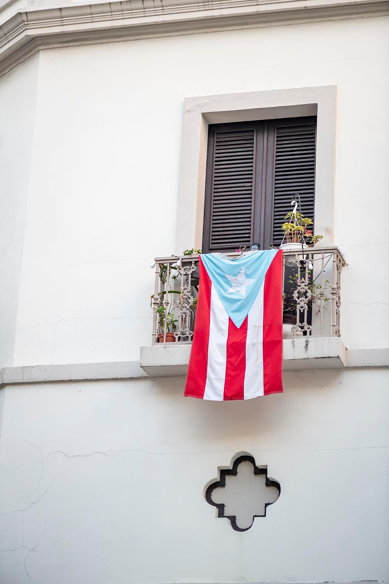 Puerto Rico flag hanging from an ornate balcony on a white building with dark shuttered windows.