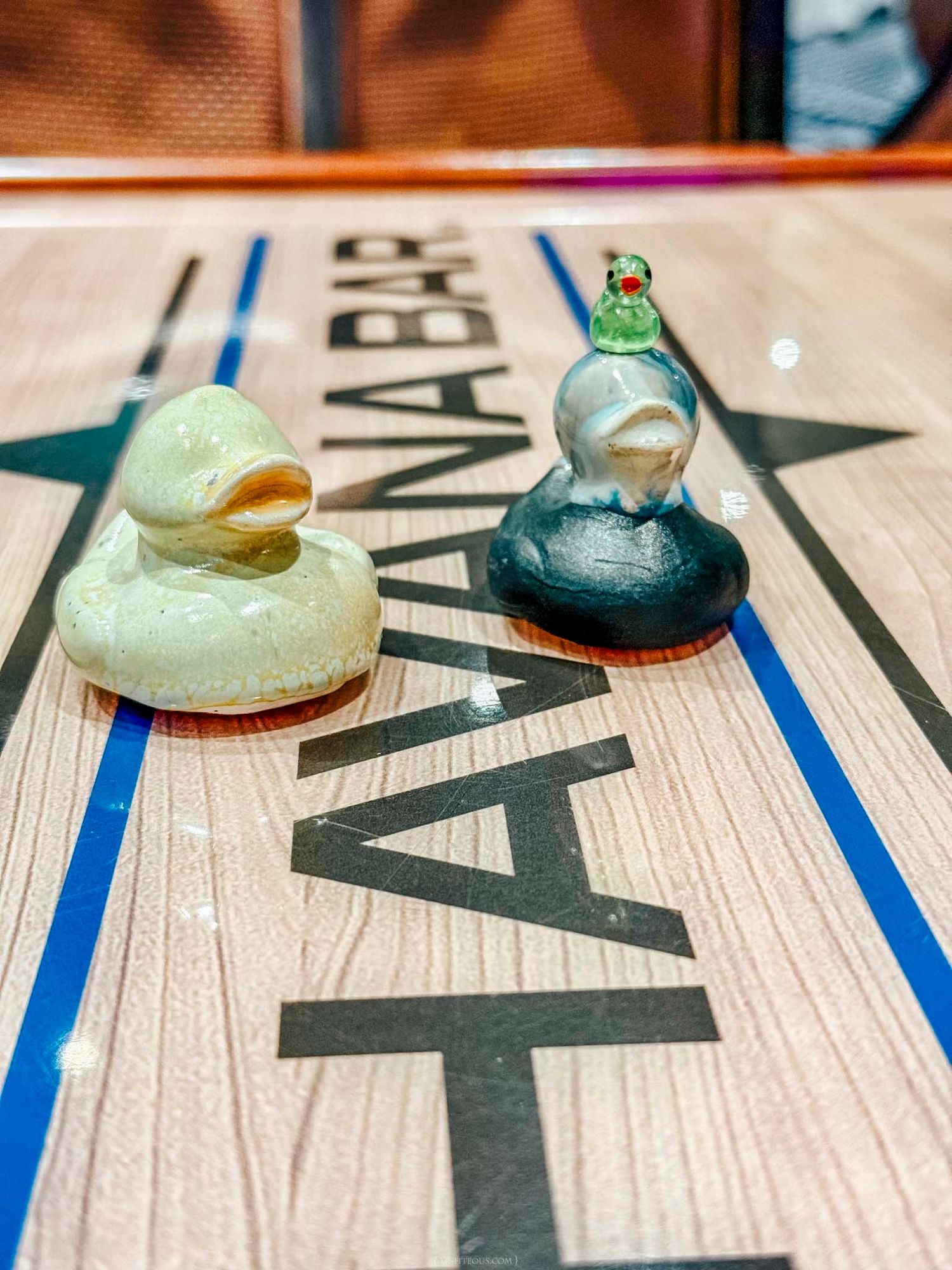 Two glass duck figurines on a shuffleboard table at a bar, one yellow and one dark with a green bird on top.