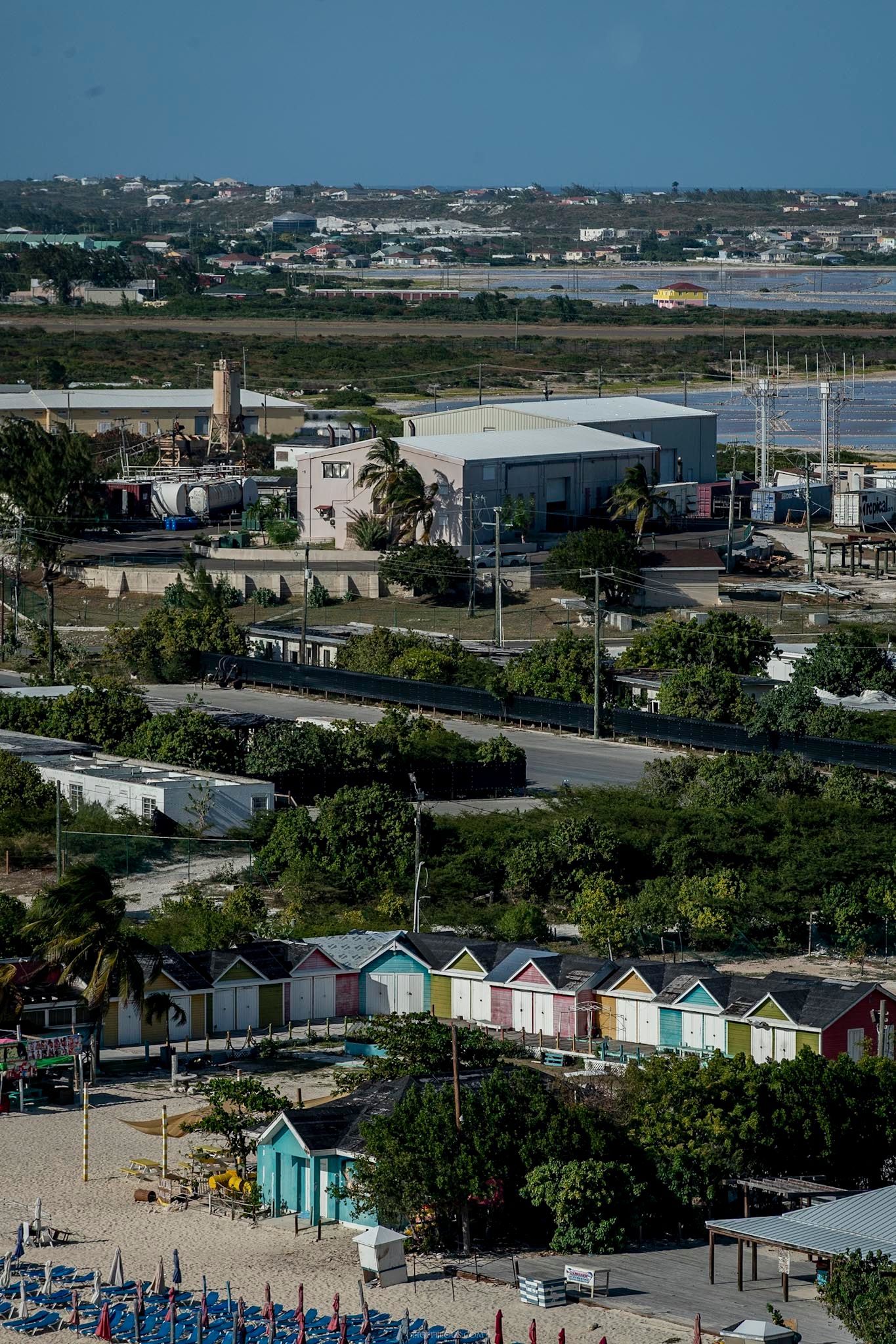 Colorful beach huts line a sandy shore near an industrial area and road in a tropical coastal town.