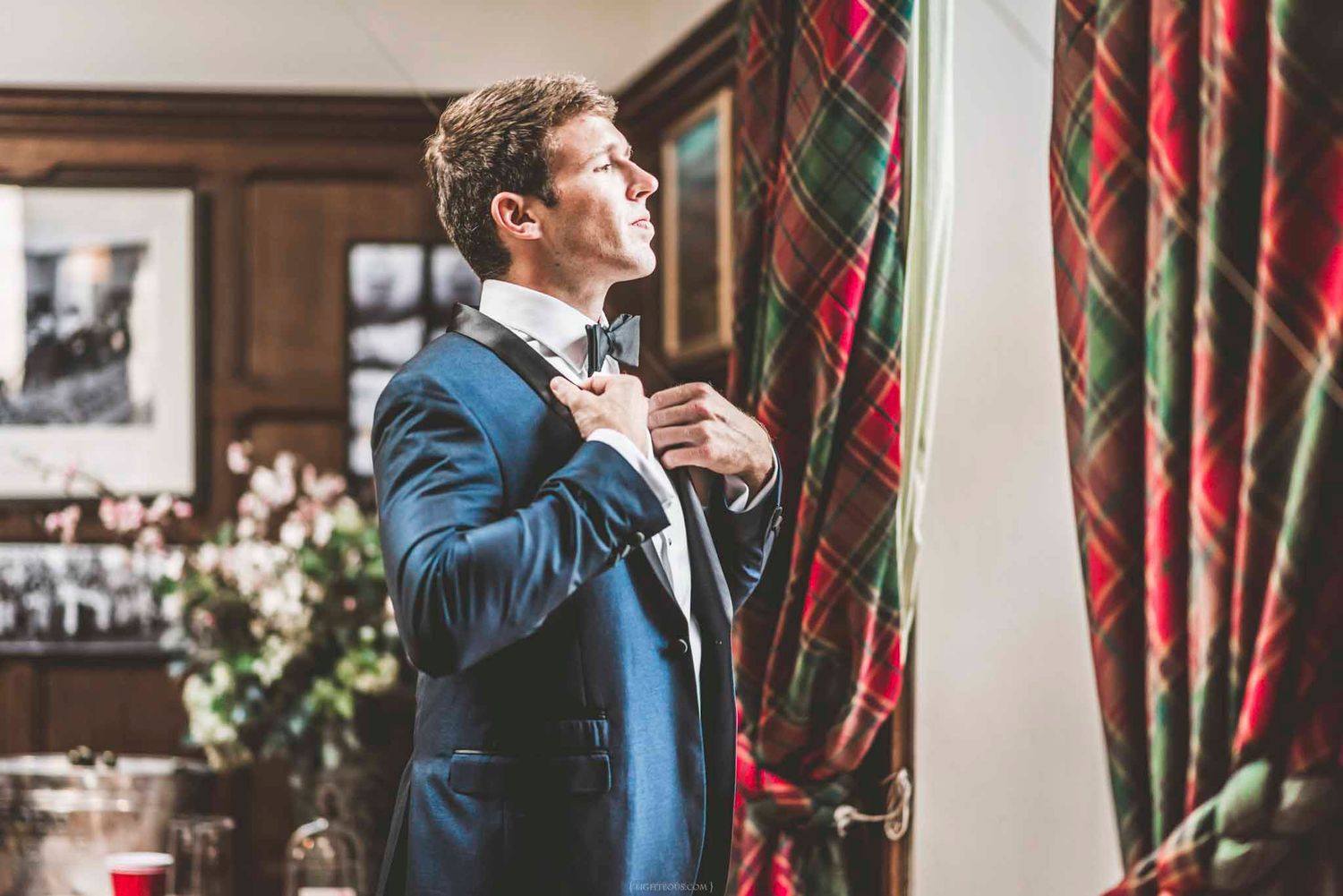 A groom in a navy tuxedo adjusts his bow tie beside plaid curtains in an elegant room.