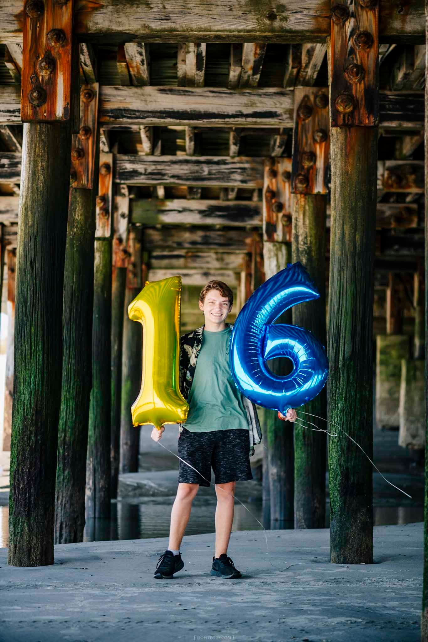 Teen boy holding gold and blue number 16 balloons under a wooden pier, celebrating his 16th birthday.