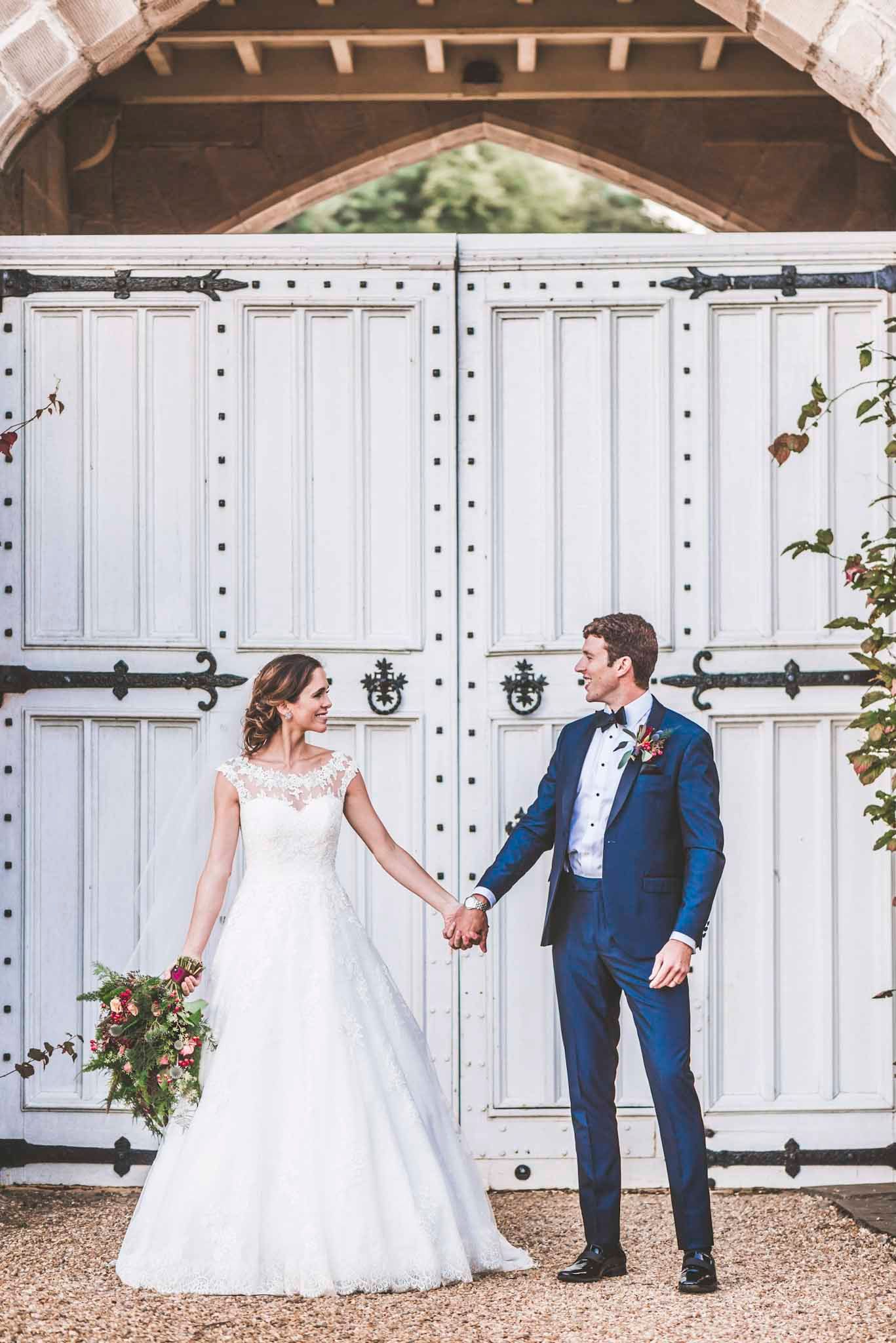 Bride and groom holding hands before white wooden gates, she in lace gown, he in navy suit with floral boutonniere.
