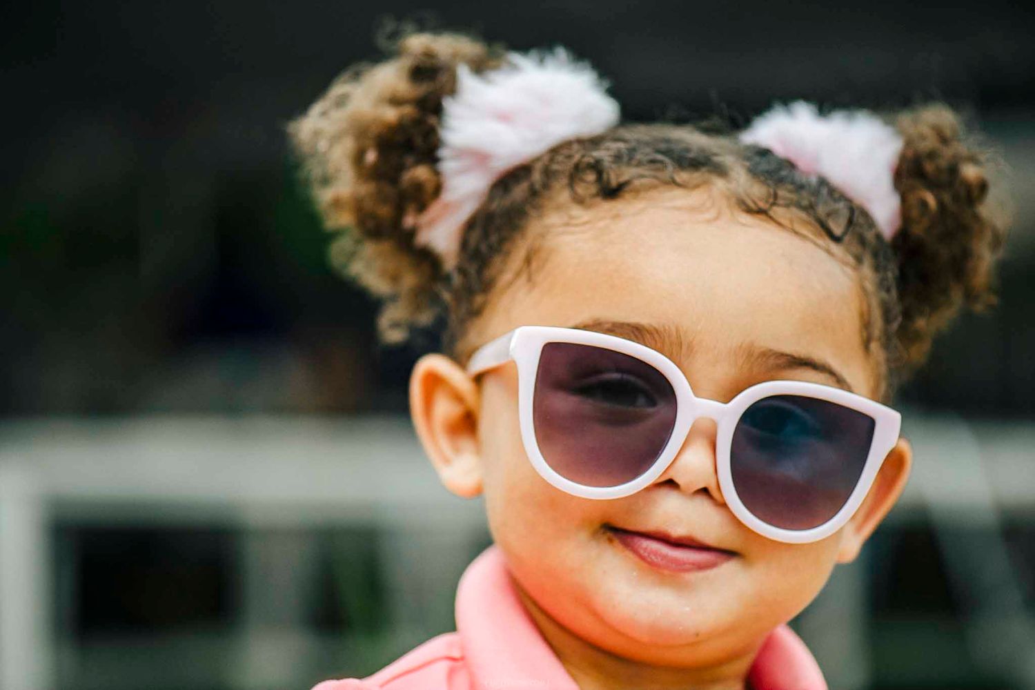 Adorable young girl with curly pigtails wearing oversized white sunglasses and a pink outfit, smiling confidently.