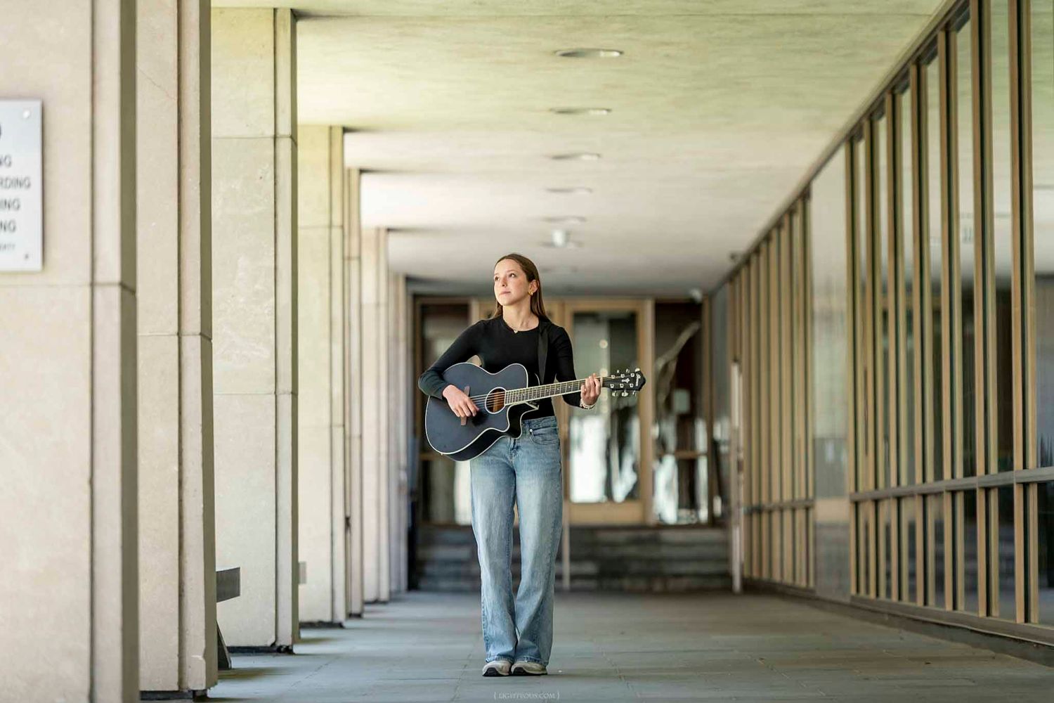 Young woman playing acoustic guitar while walking through a modern covered outdoor corridor.