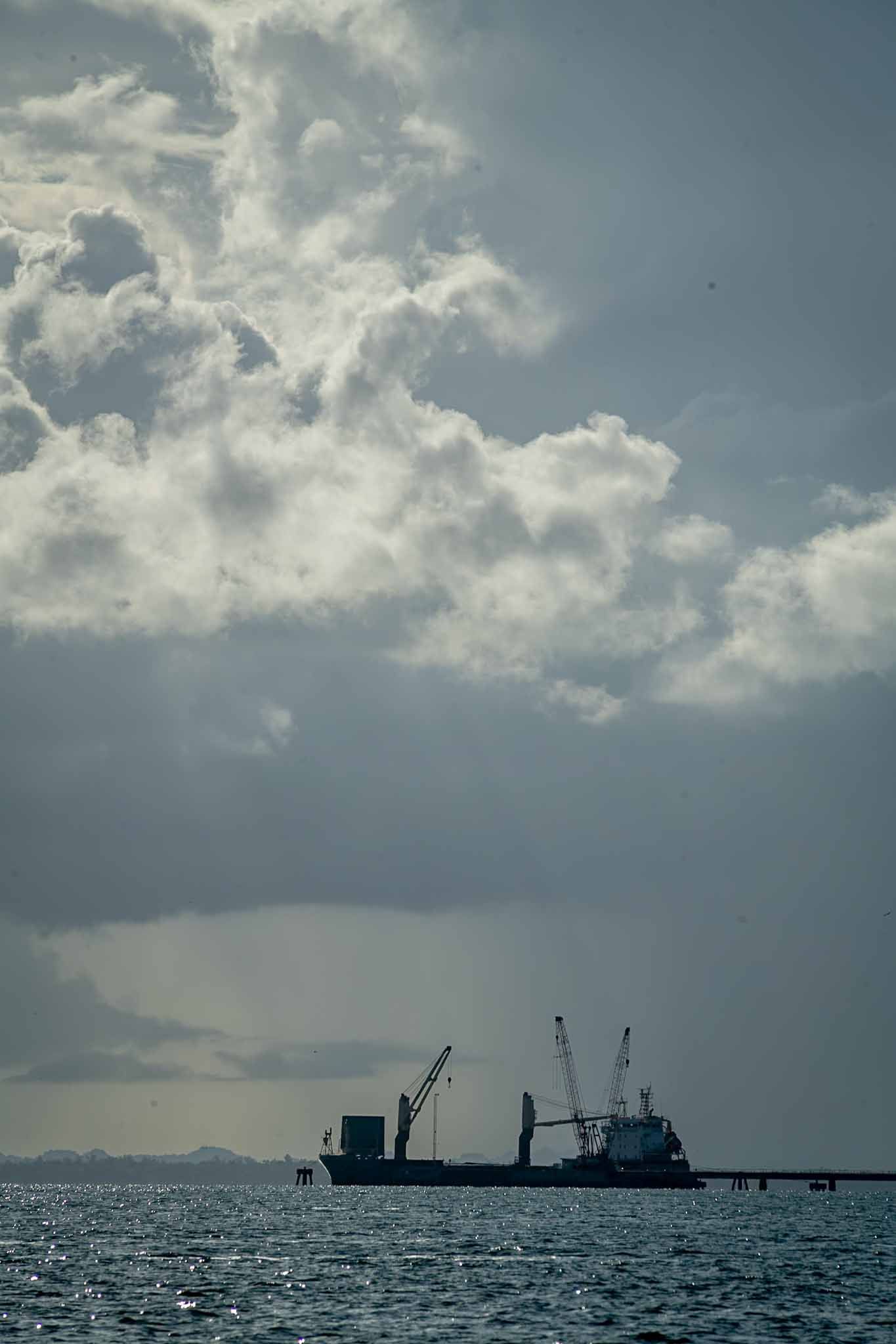 Silhouette of cargo ship with cranes against dramatic cloudy sky over dark ocean waters.