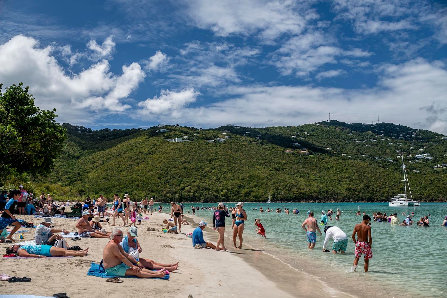 Tourists enjoy a sunny Caribbean beach with turquoise waters and lush green hills in the background.