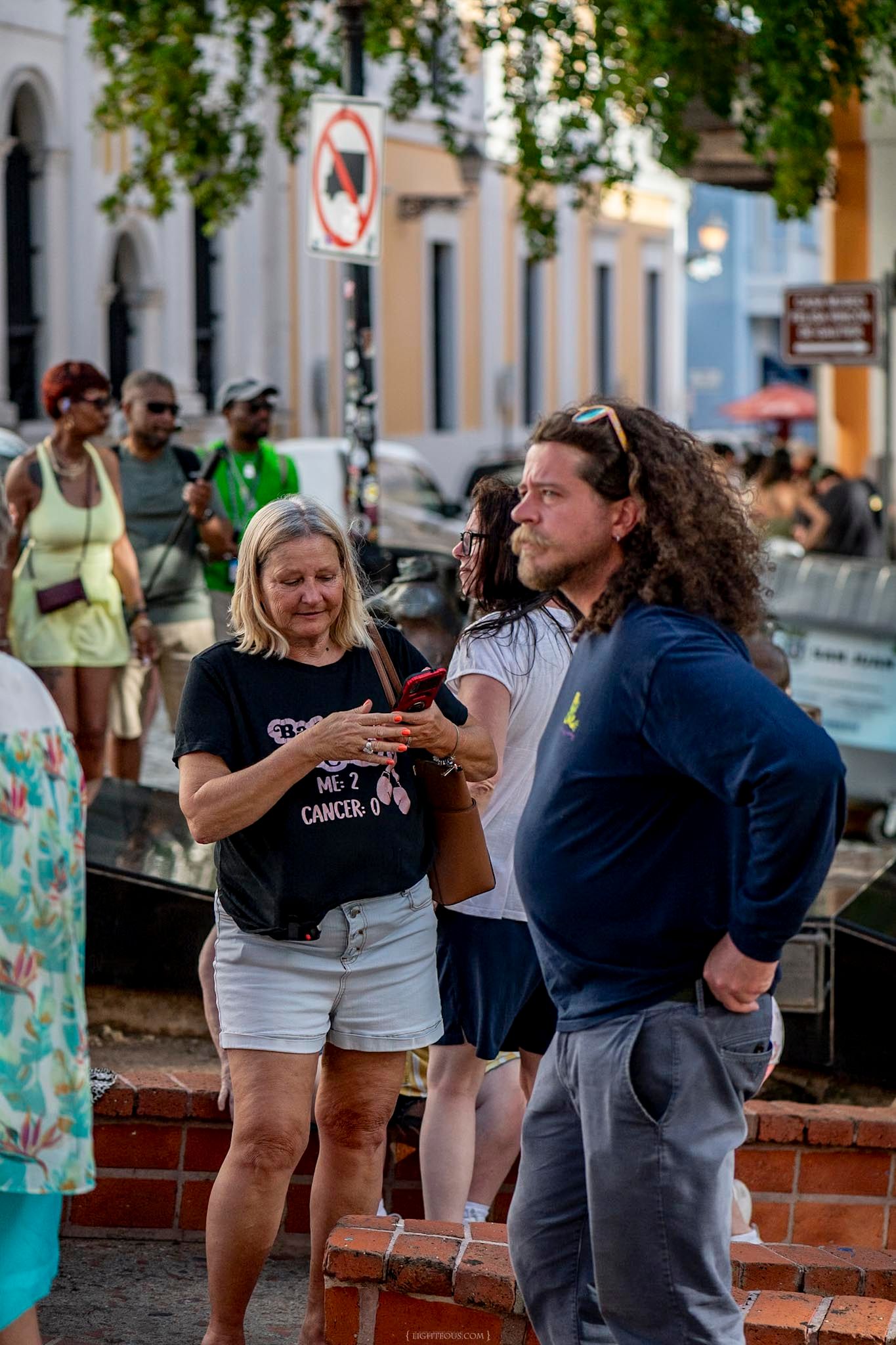 Tourists gather on a busy urban street, with a woman wearing a cancer awareness shirt checking her phone.