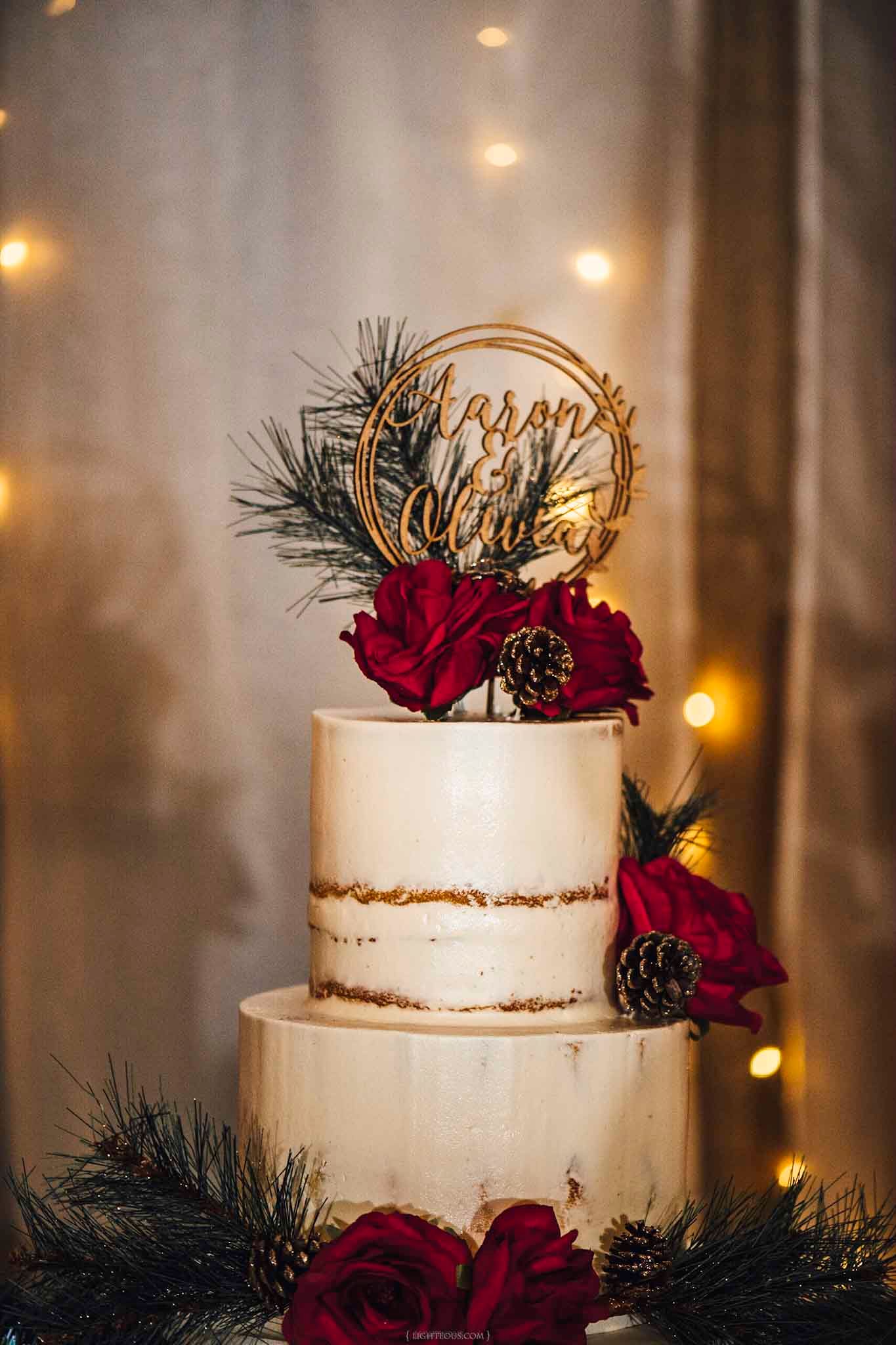 Two-tier naked wedding cake with red roses, pine cones, and gold personalized topper surrounded by fairy lights.