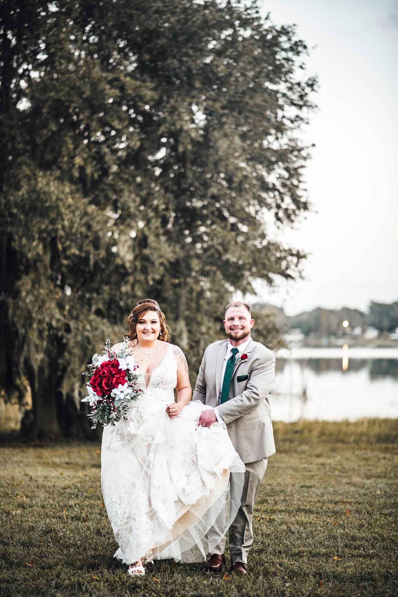 Bride in lace gown holding red bouquet with groom in gray suit by a lakeside at dusk.