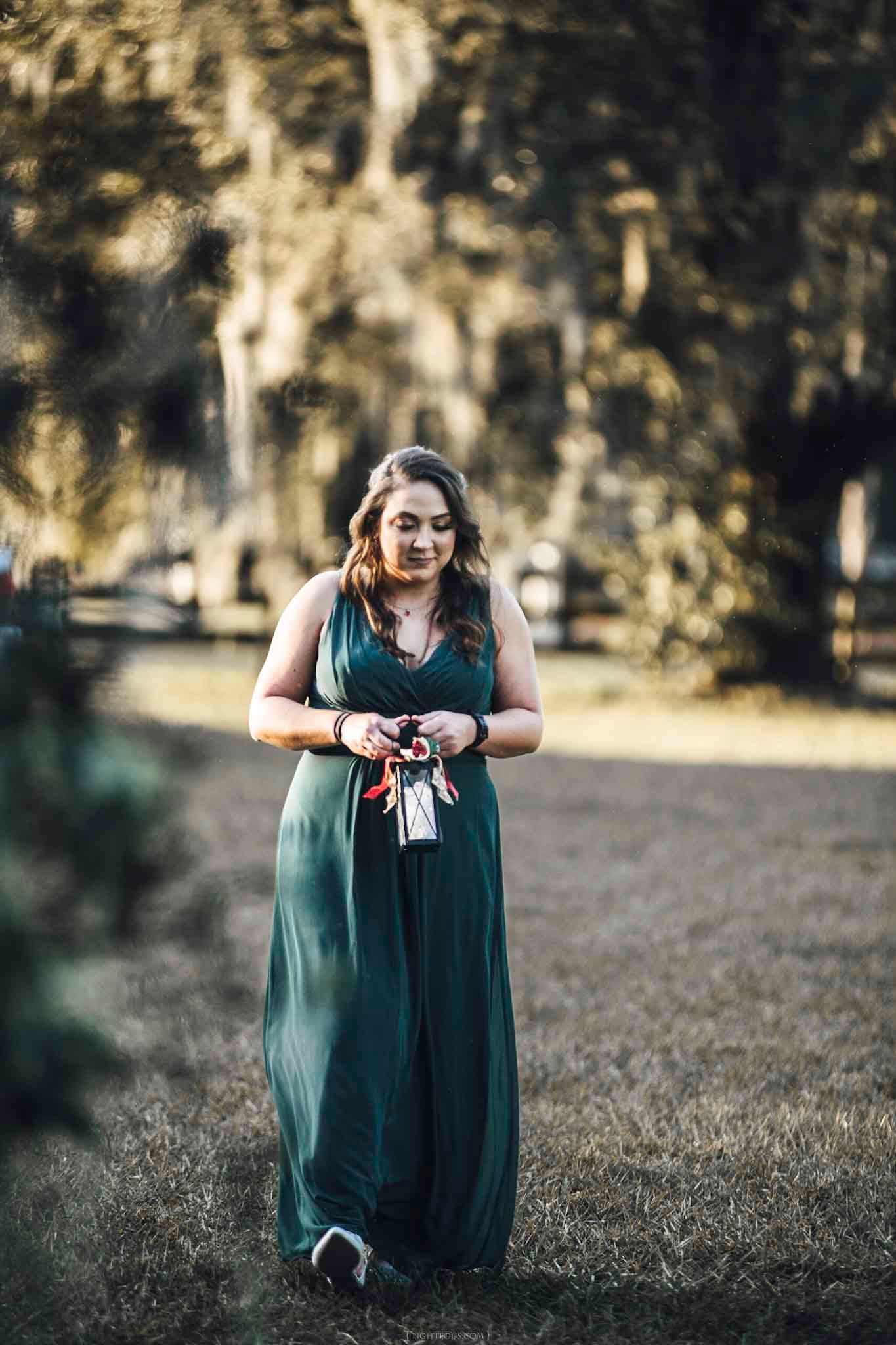 Woman in elegant green maxi dress standing outdoors among moss-draped trees in a serene garden setting.
