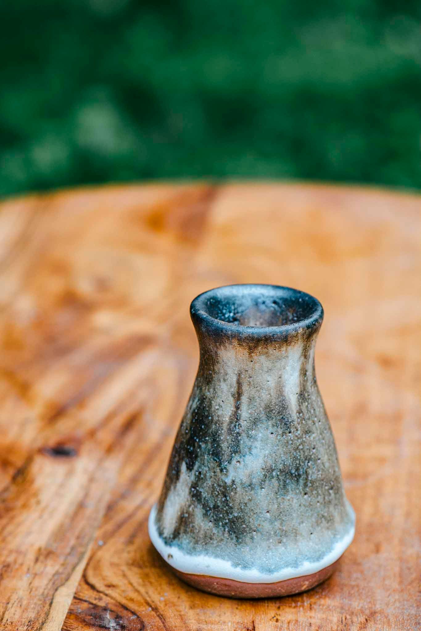 Small handcrafted ceramic vase with blue-gray glaze on a rustic wooden table with green background.