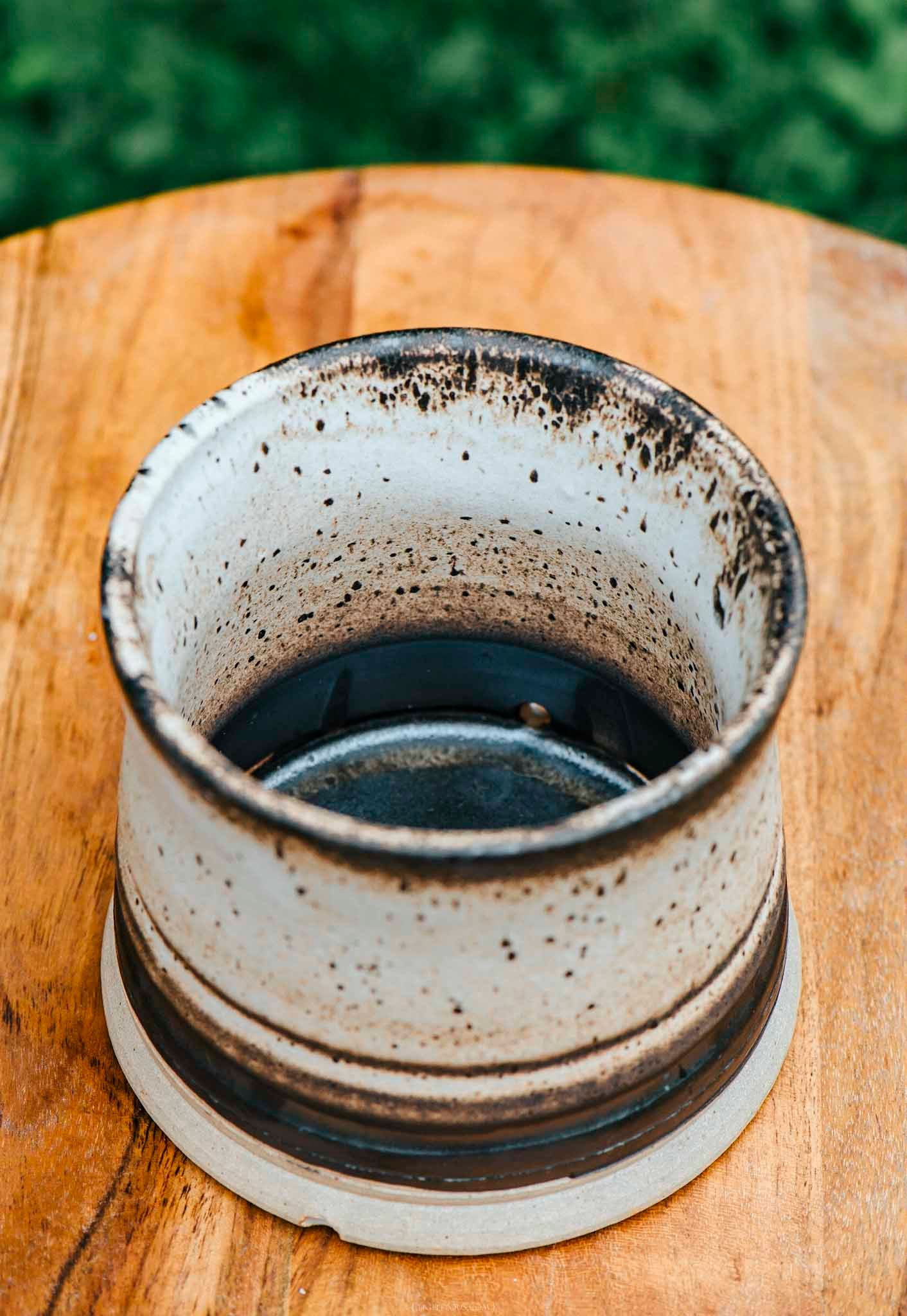 Rustic speckled ceramic cup with dark glaze interior sitting on a wooden table with green foliage background.