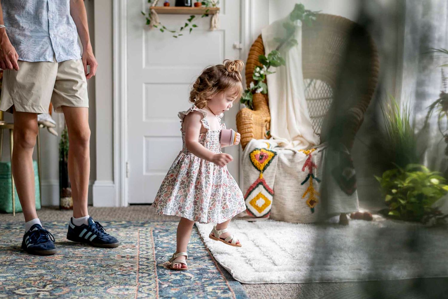A toddler girl in a floral dress walks barefoot on a patterned rug in a bright boho-styled living room.