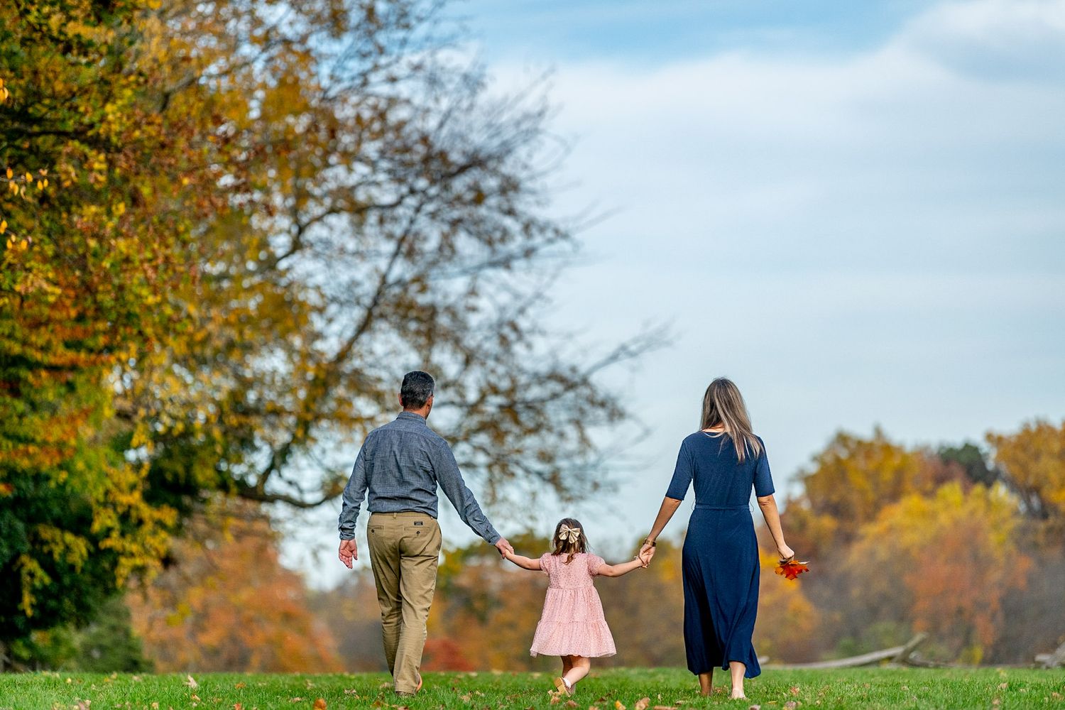 Family of three walking hand-in-hand through a lush green park surrounded by colorful autumn foliage on a sunny day.
