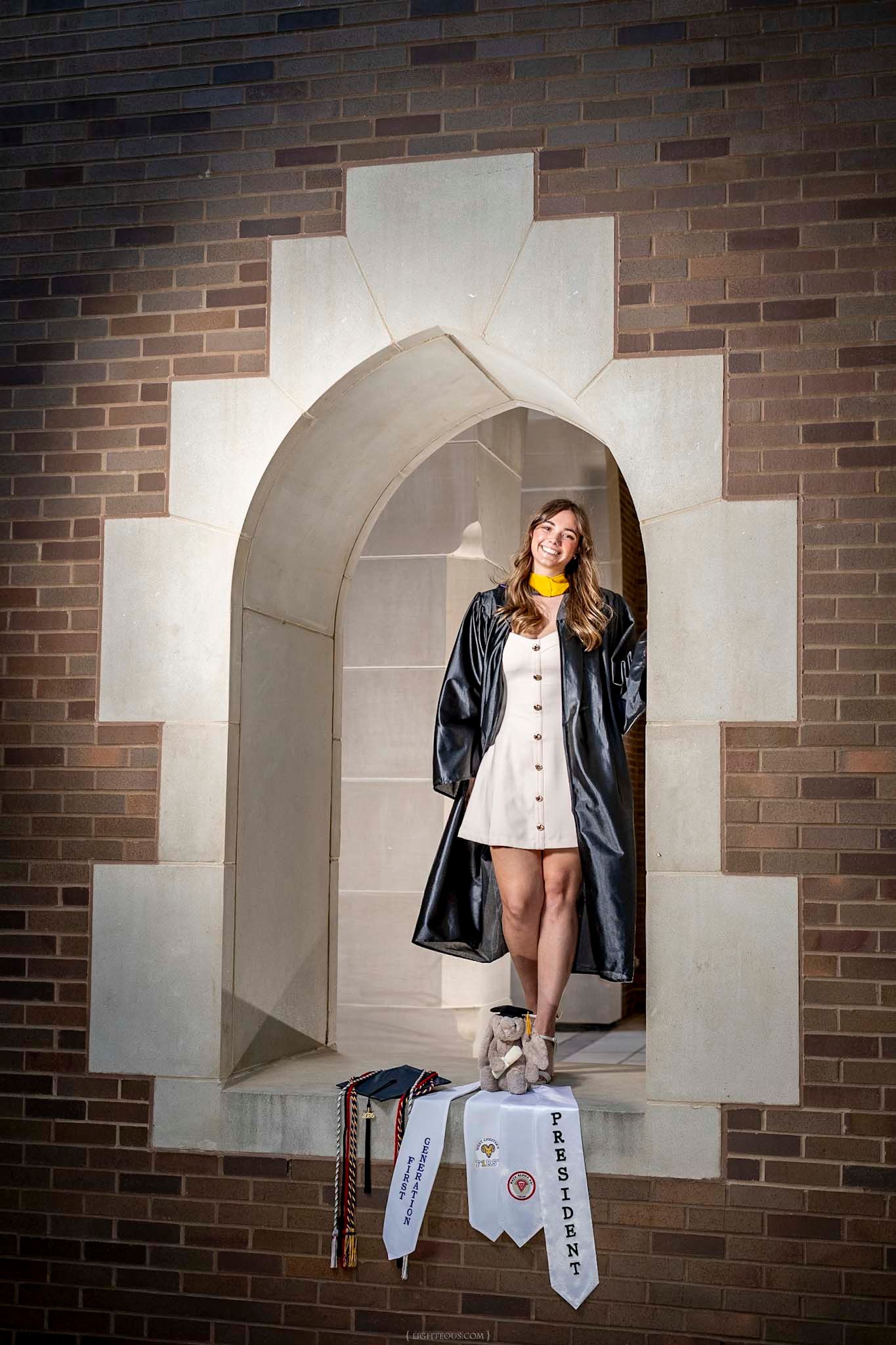 Young woman in graduation gown and white dress stands in stone archway with honor stoles, celebrating her academic achievements.