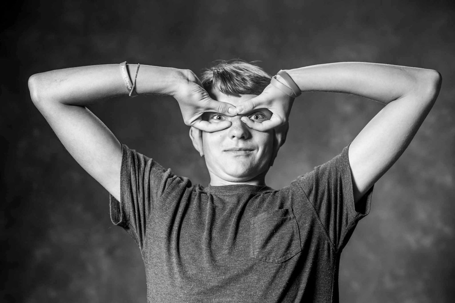 Black and white photo of a young person making funny faces with hands framing eyes against dark background.