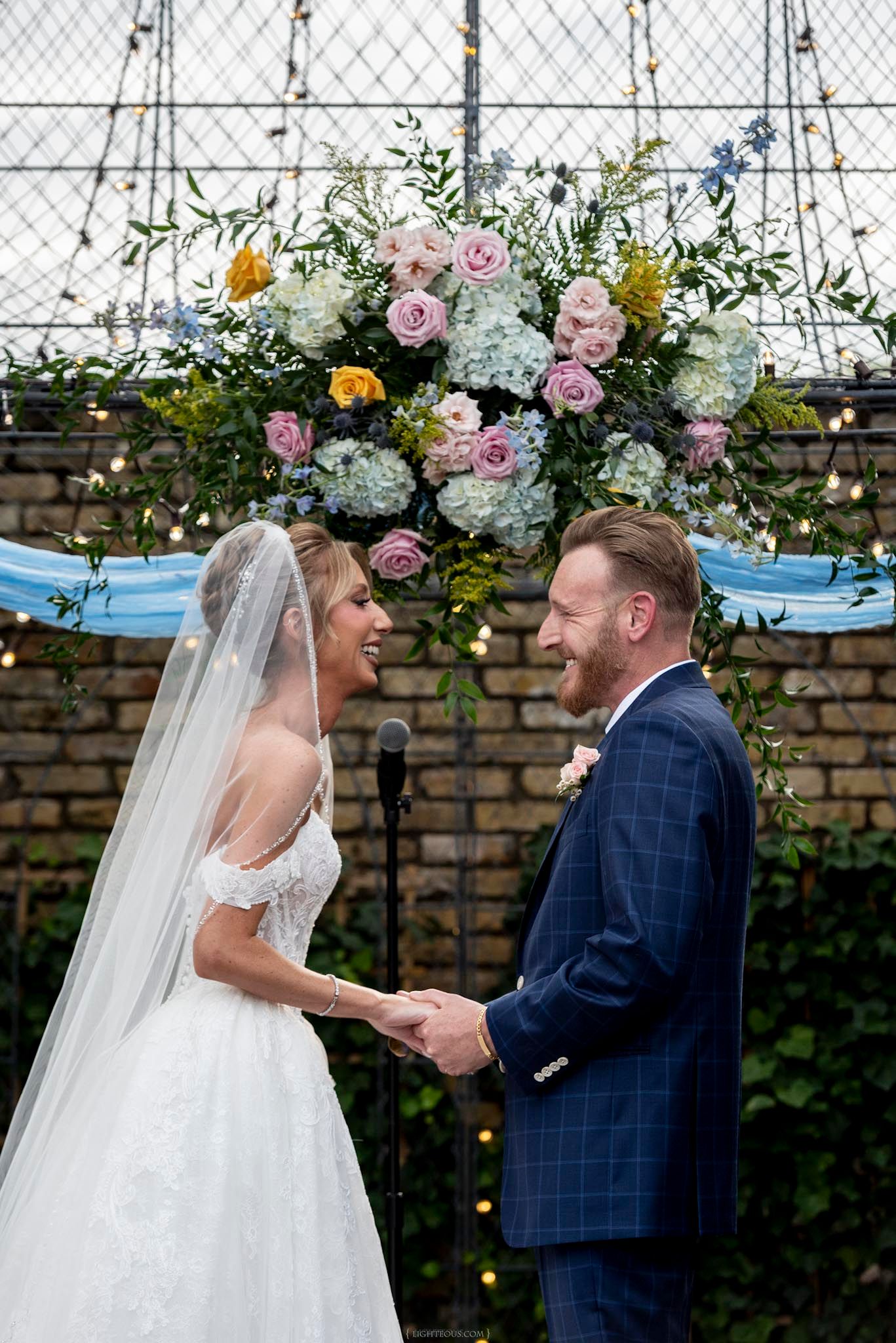 Bride and groom laughing together at outdoor wedding ceremony beneath floral arch with pink roses and blue hydrangeas.