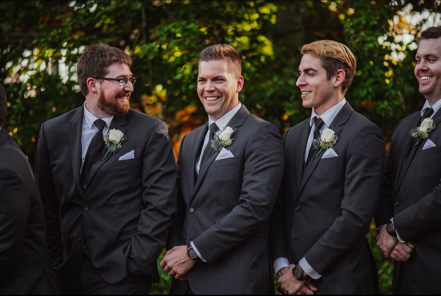 Groomsmen in charcoal suits with white boutonnieres smiling outdoors at a wedding ceremony.