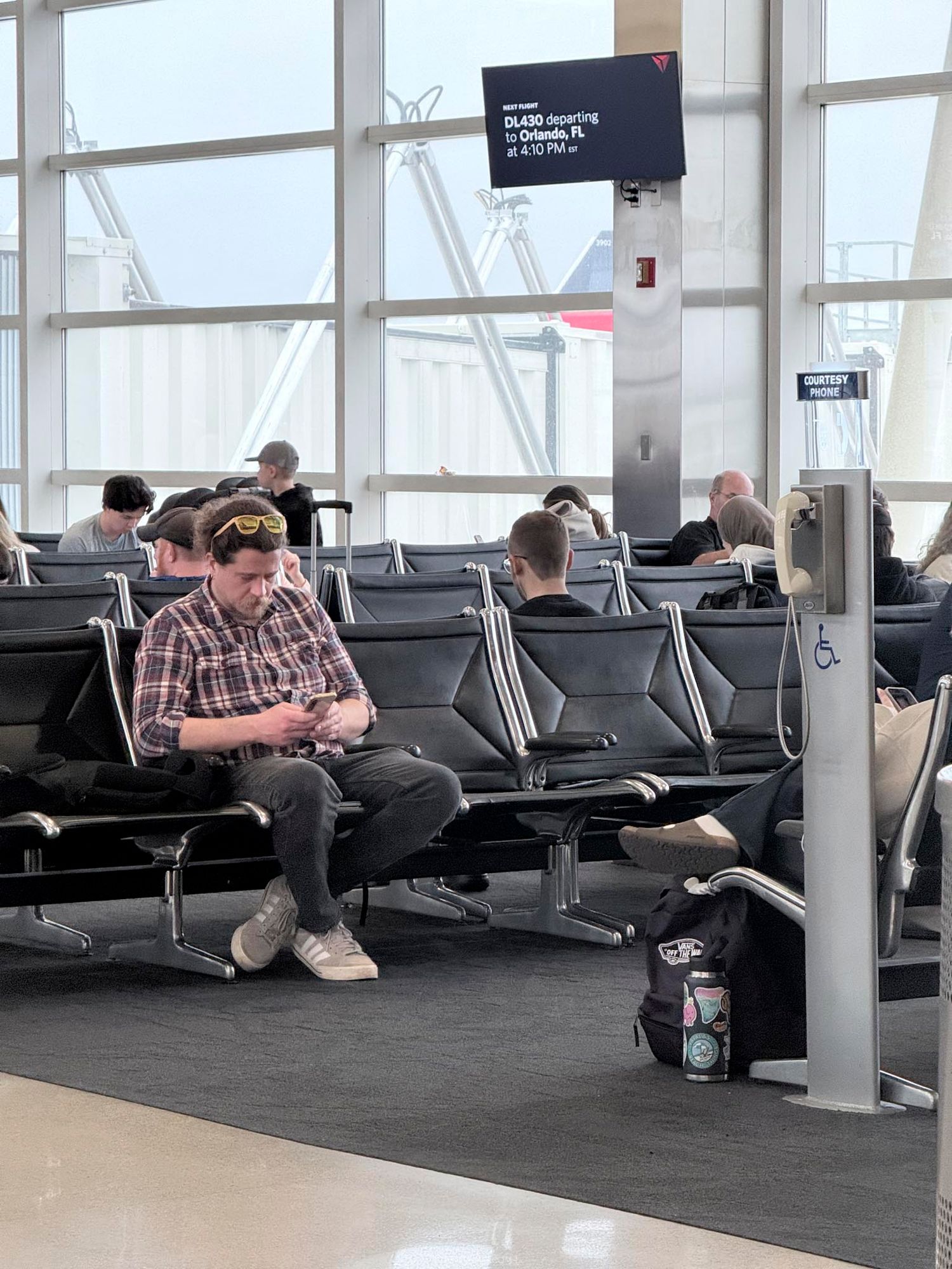 Man in plaid shirt uses phone at airport gate seating area with departure board visible overhead.