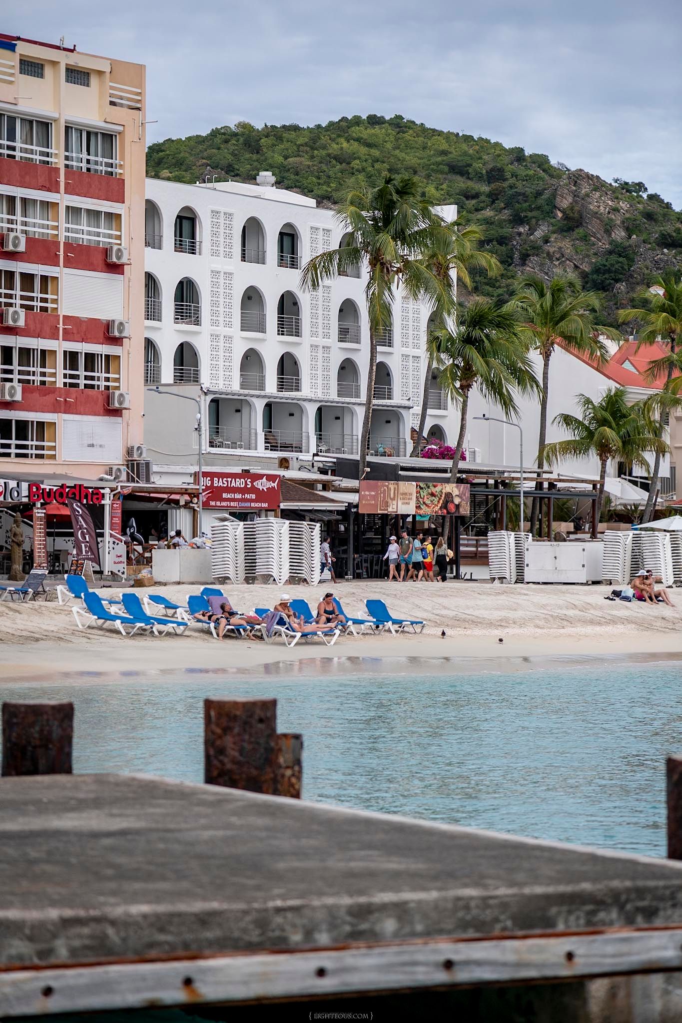 Tropical beach resort with blue lounge chairs, palm trees, white buildings and a green hillside in St. Maarten.