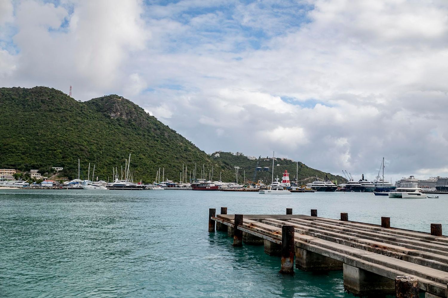 A wooden dock overlooks a scenic harbor with sailboats and a lush green hillside under a cloudy sky.