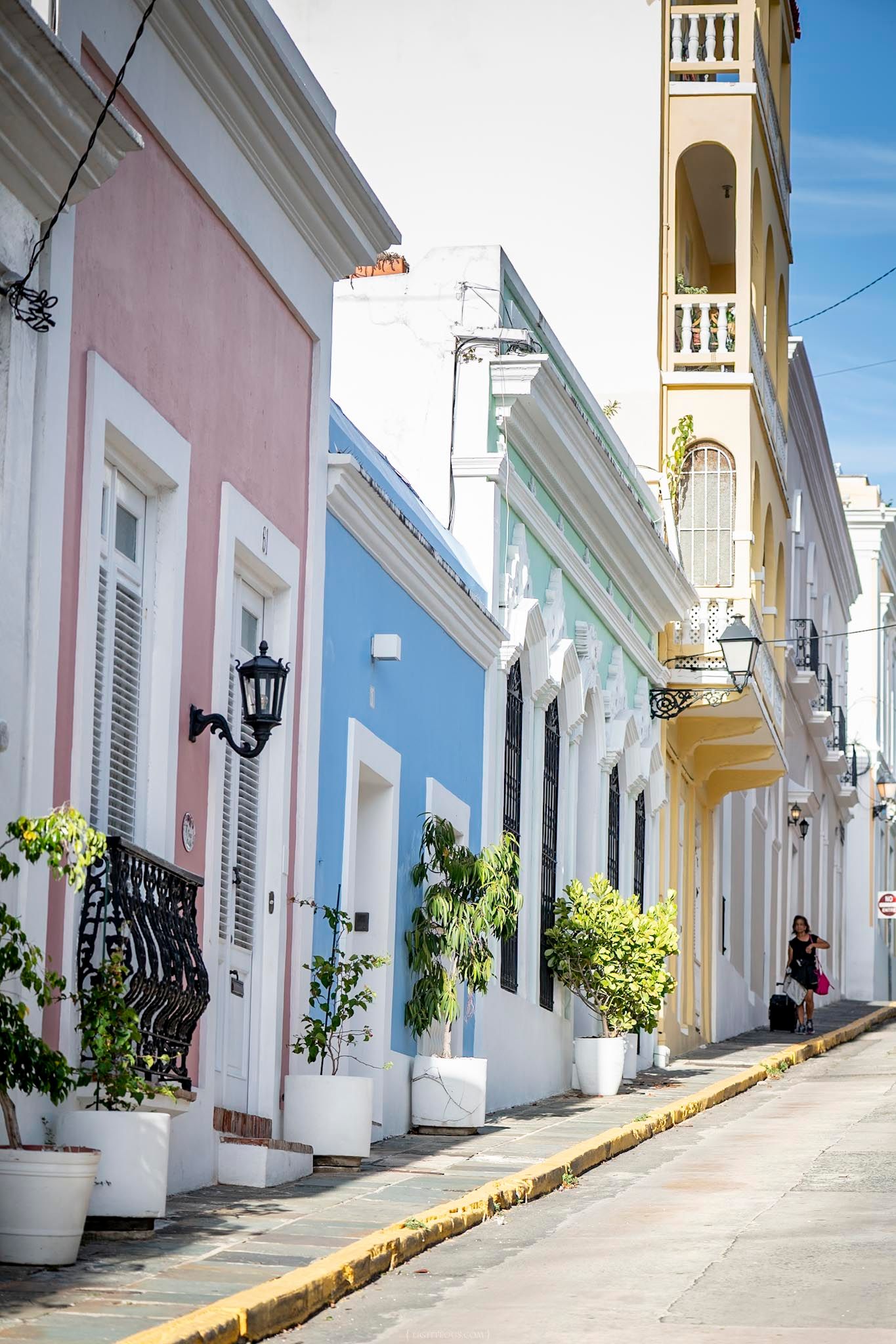 Colorful colonial buildings line a sunny cobblestone street with potted plants and ornate balconies in Old San Juan.