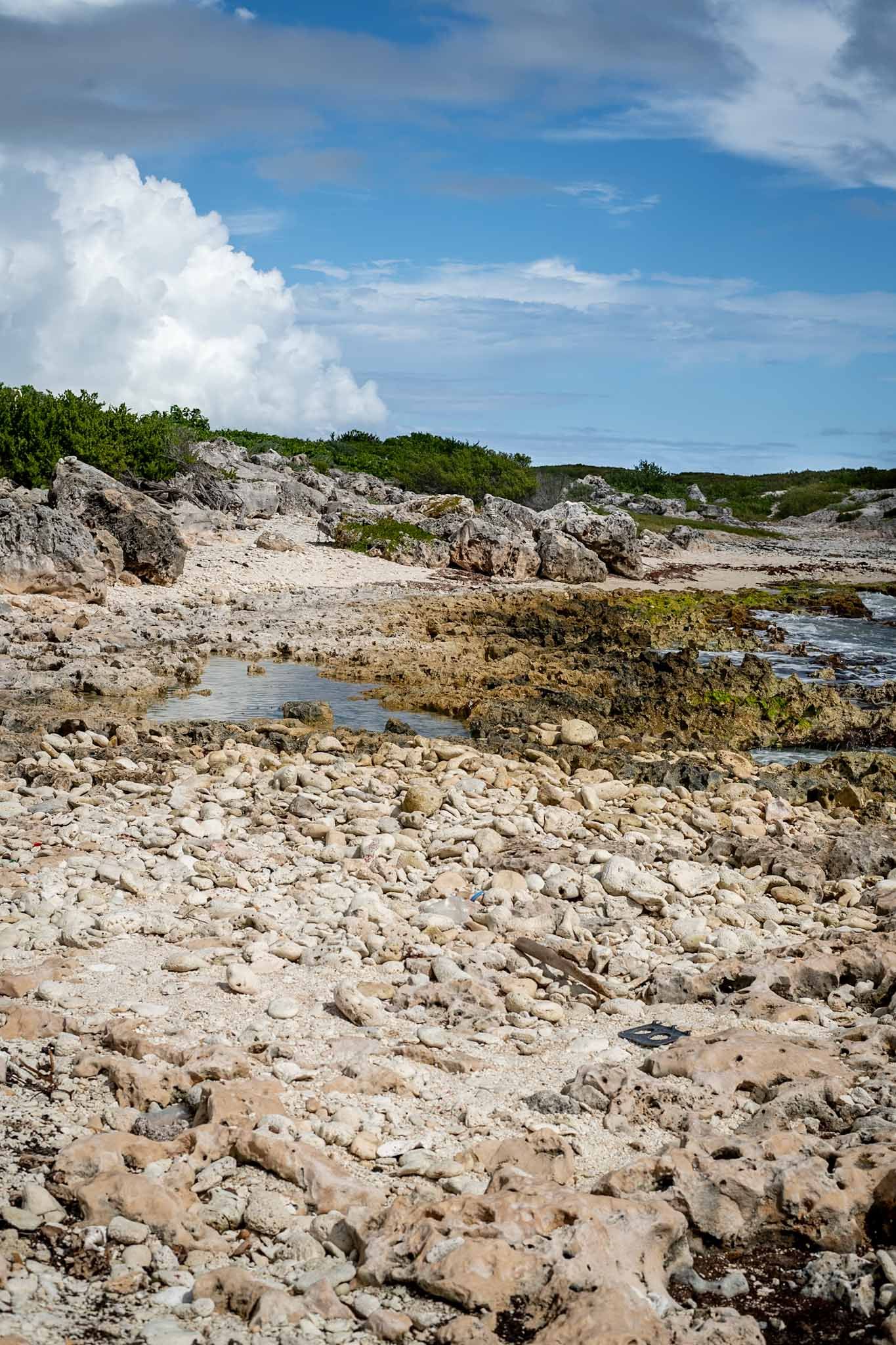 Rocky limestone coastline with turquoise water pools and white clouds in blue sky over tropical beach landscape.