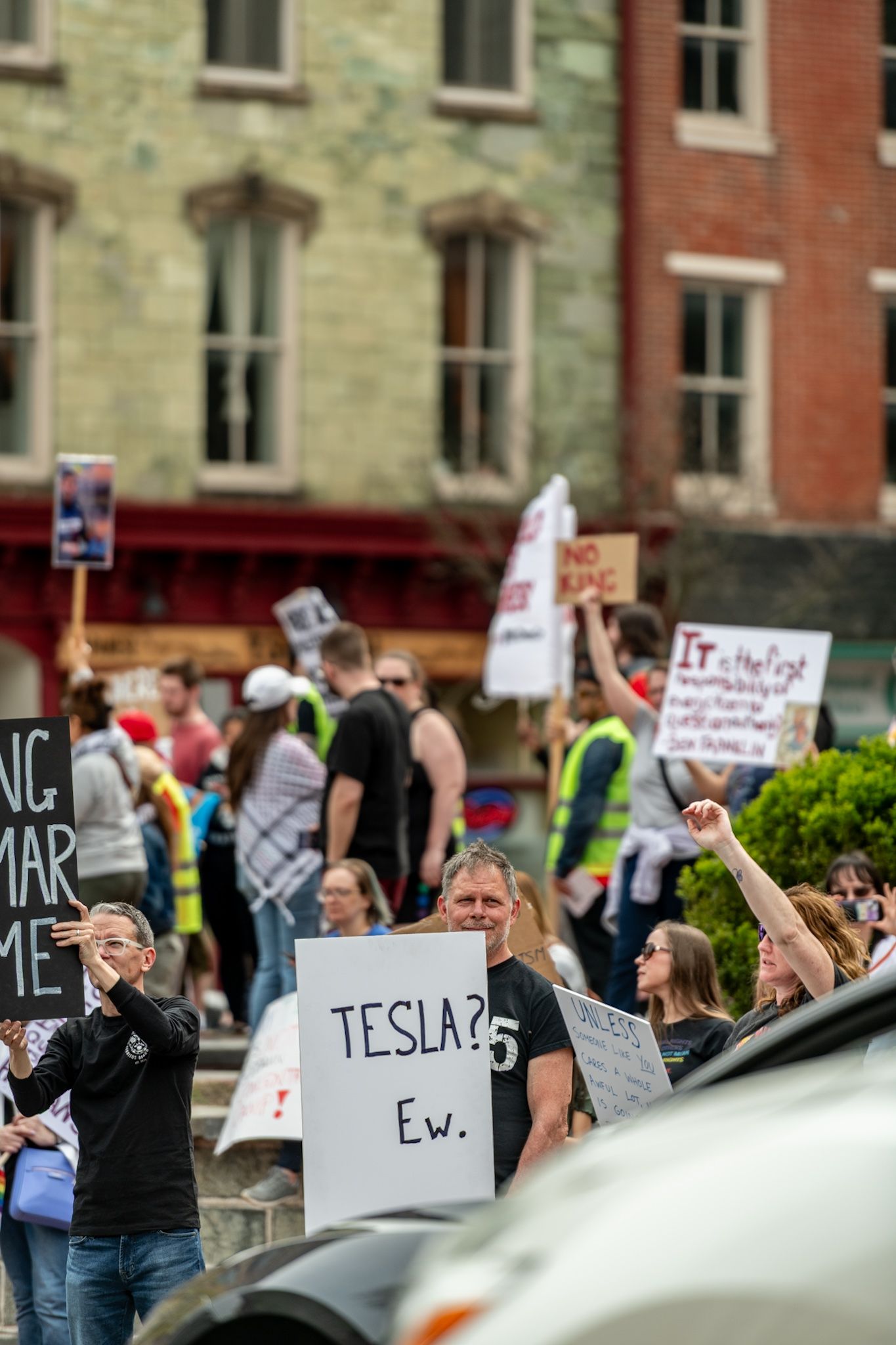 Protesters with signs gather along a street with historic stone buildings in the background.