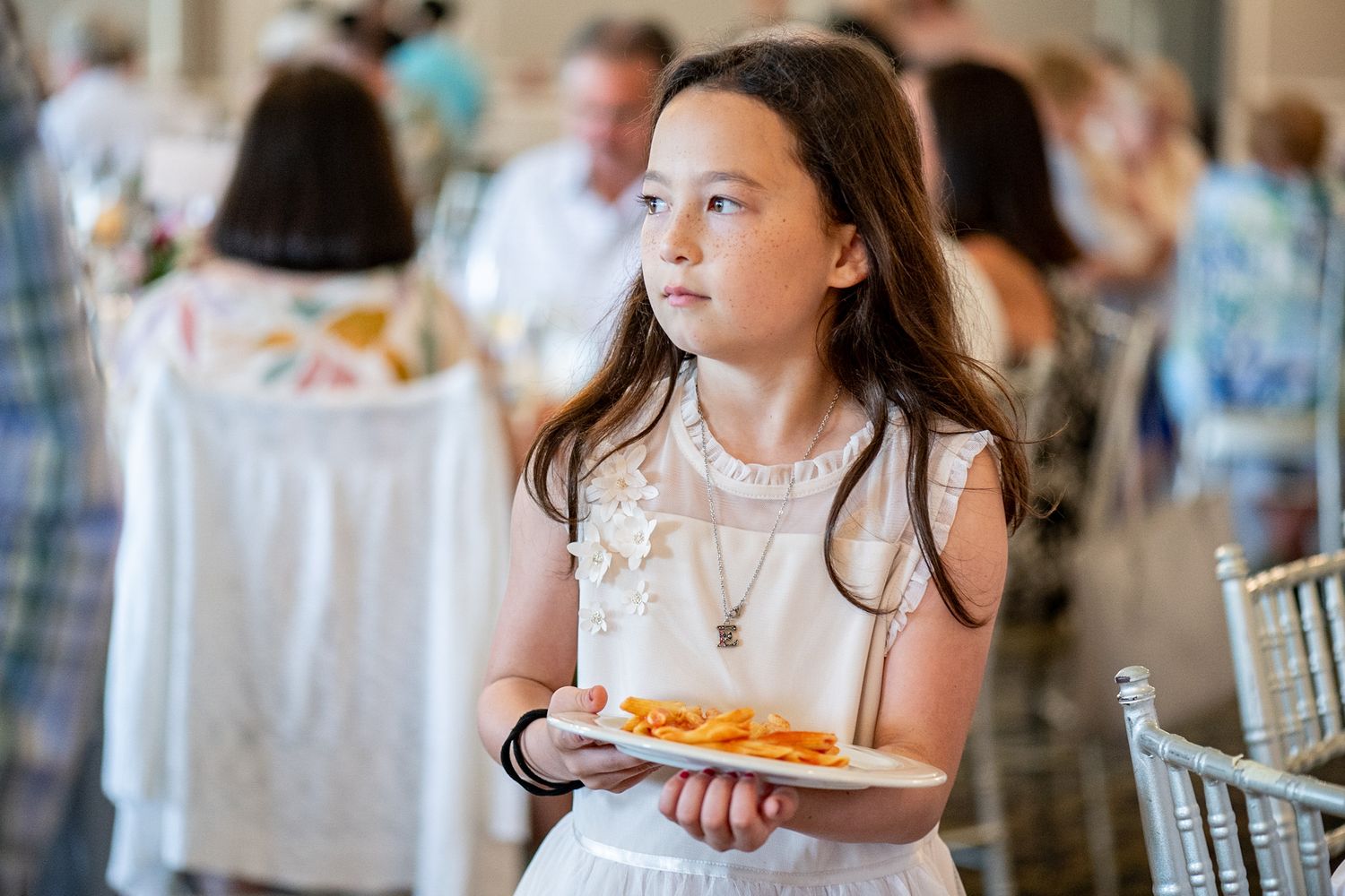 Young girl in white dress holding a plate of food at an elegant indoor party or celebration.