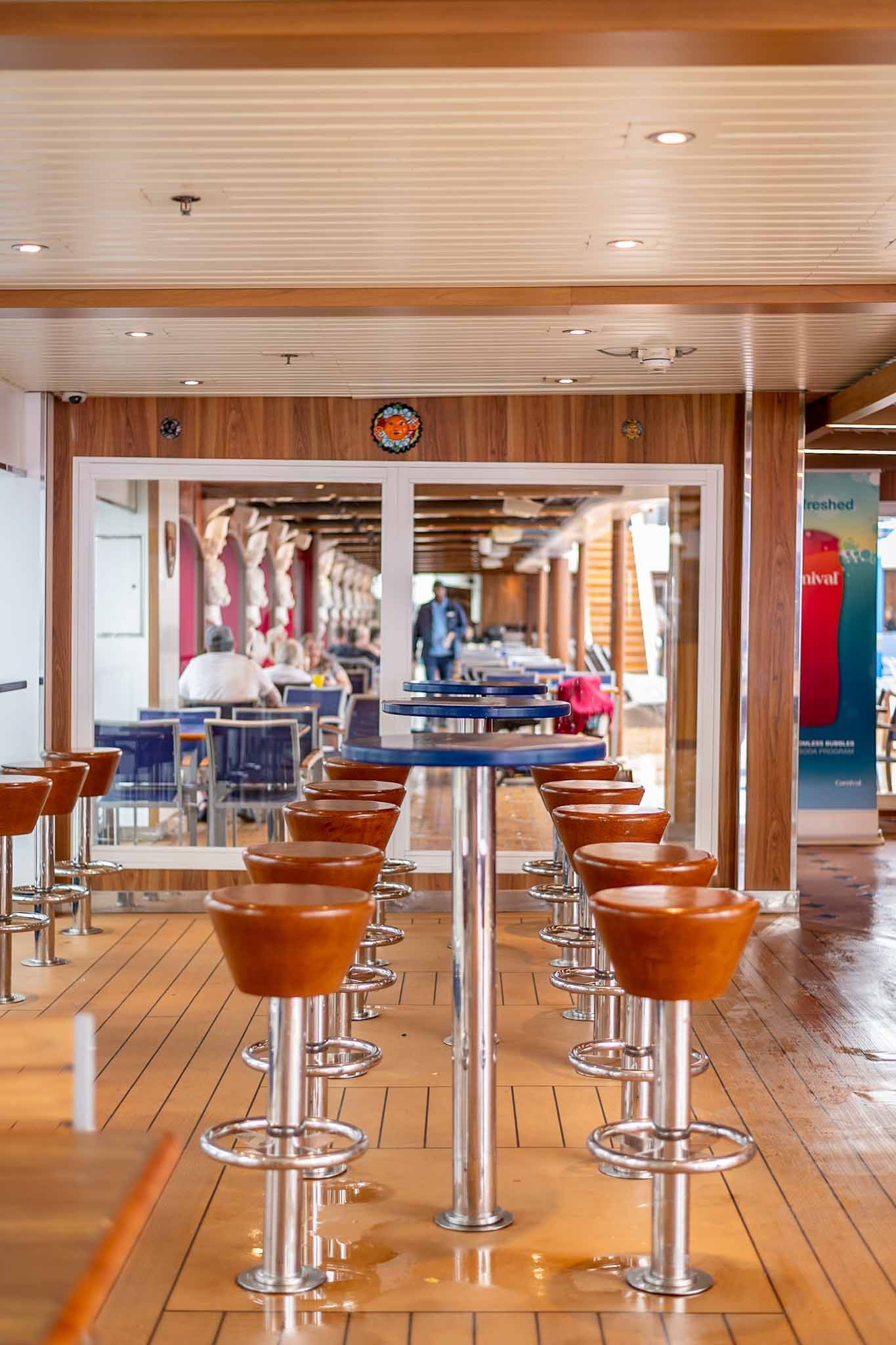 Modern cruise ship bar area with wooden deck flooring and sleek chrome bar stools arranged in rows.