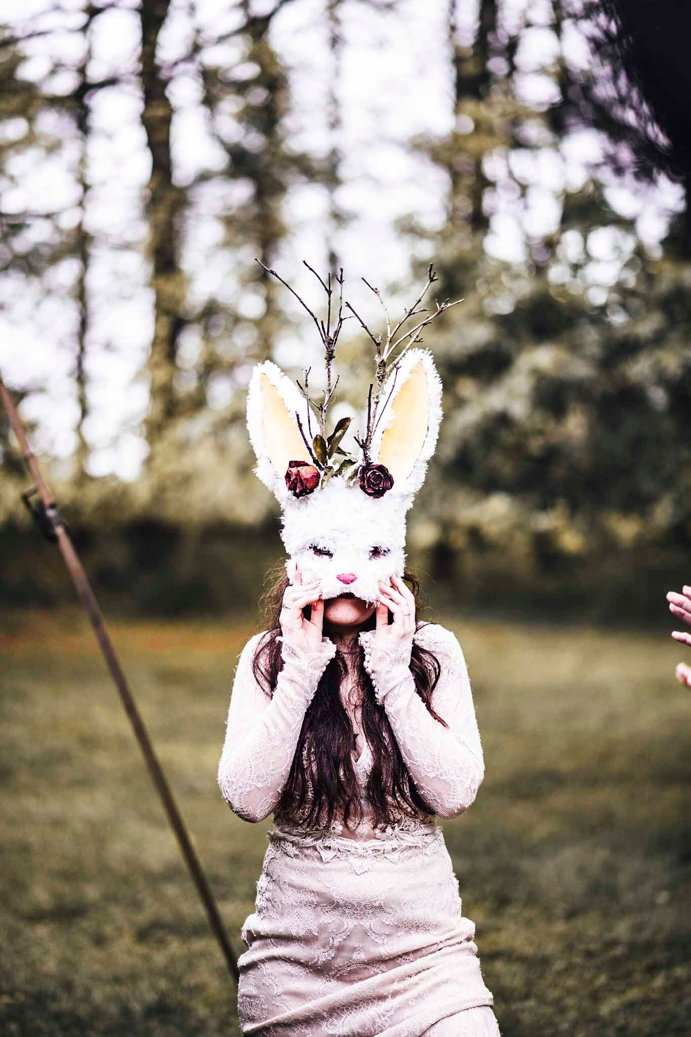 Person wearing a white bunny mask with floral decorations and twigs, standing outdoors in a forest setting.