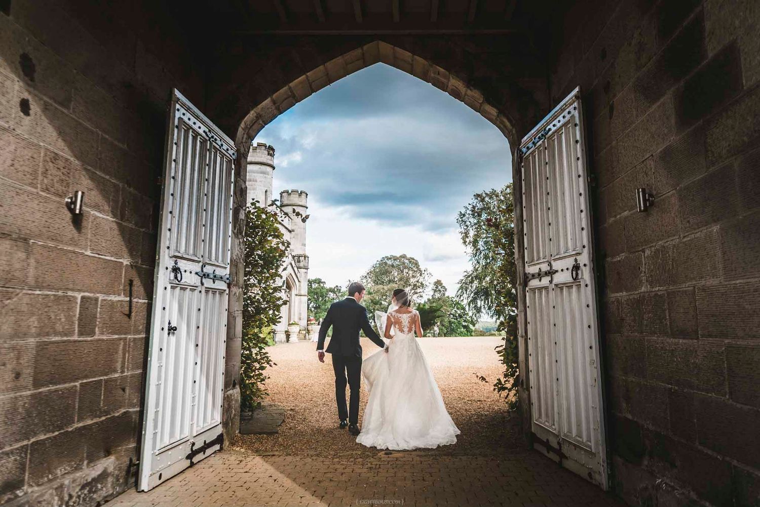 Bride and groom walk hand in hand through a grand stone archway toward a castle at their wedding venue.