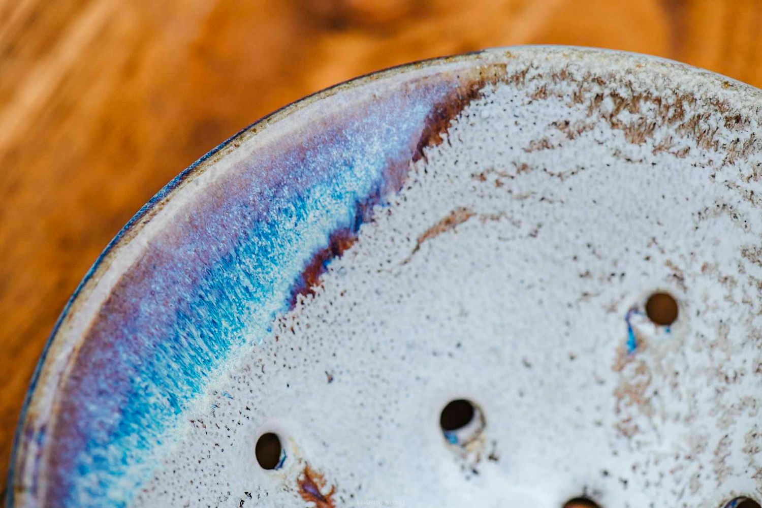 Close-up of a handmade ceramic soap dish with blue and white glaze, featuring drainage holes on a wooden surface.
