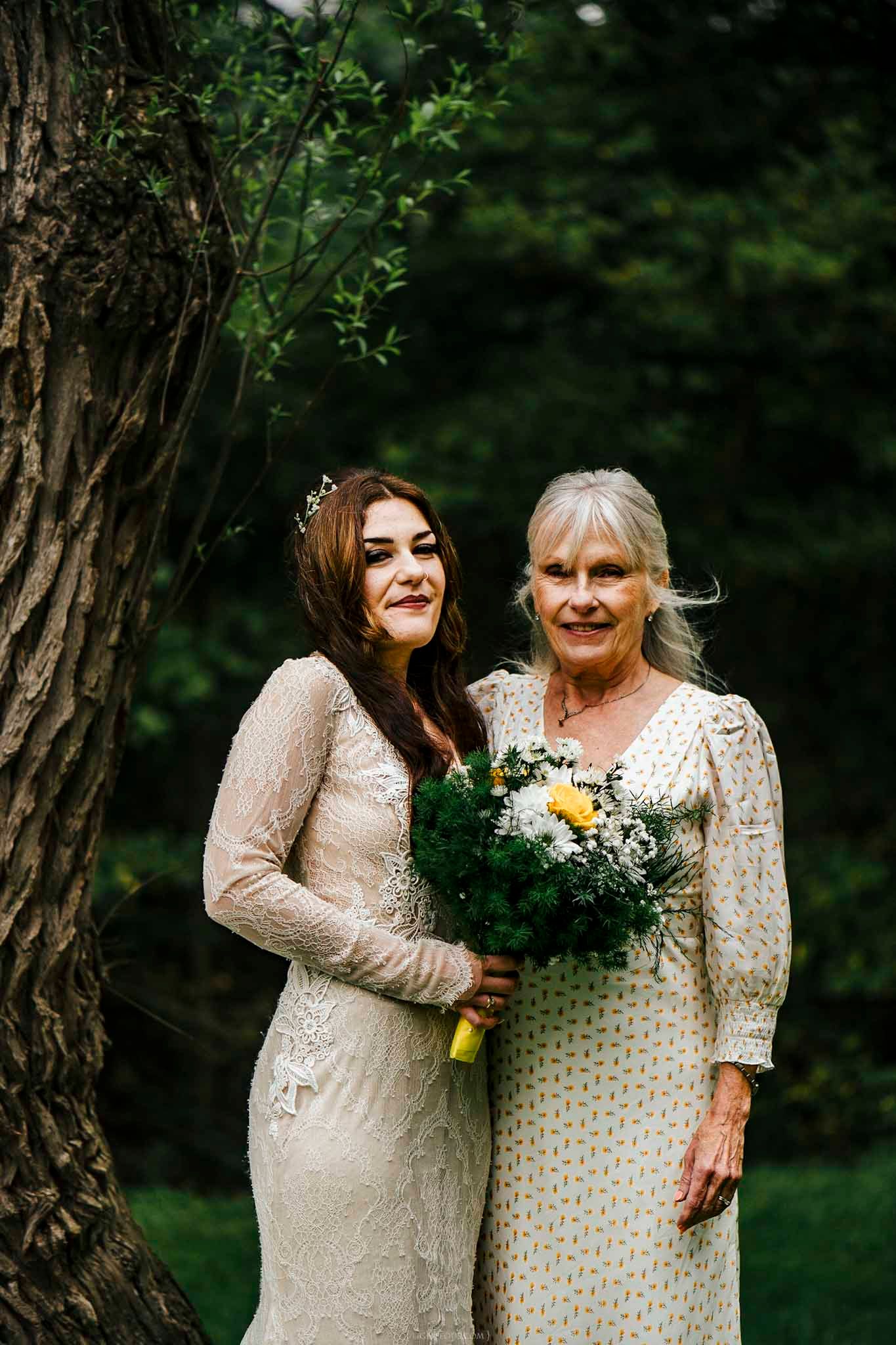Bride in lace gown and older woman in floral dress holding bouquet pose together outdoors near a tree.