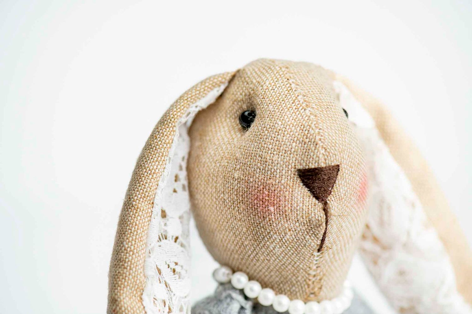 Close-up of a handmade burlap stuffed bunny toy with lace collar and pearl necklace on white background.