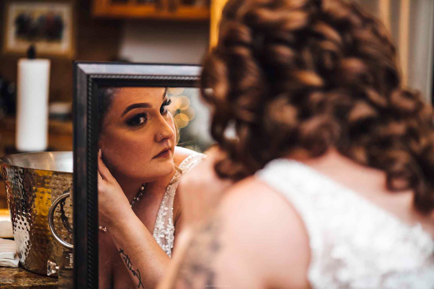 Bride in lace wedding dress examines her makeup and hair in a mirror while getting ready on her wedding day.
