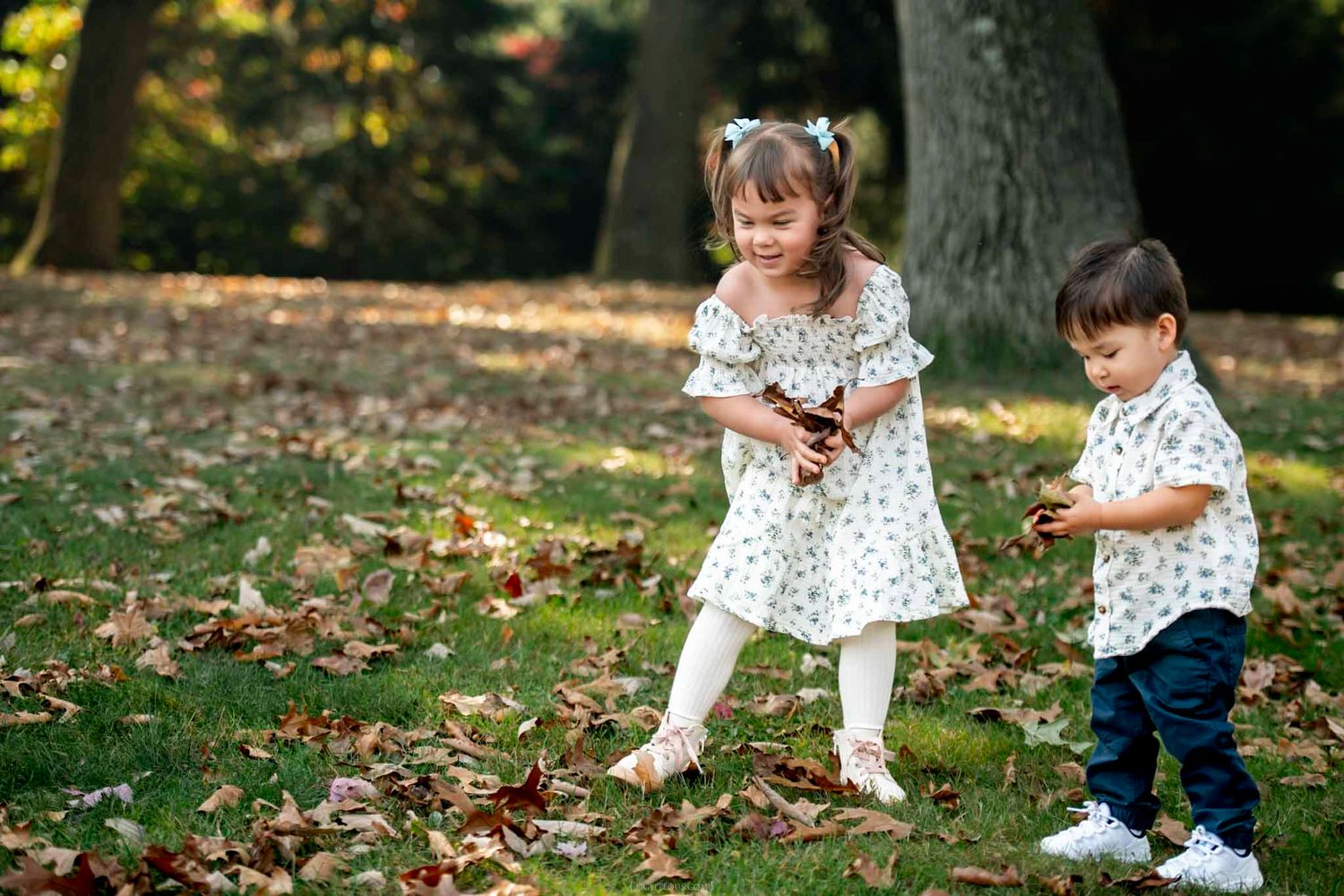 Two young children playing with autumn leaves in a park, girl in white dress and boy in white shirt.