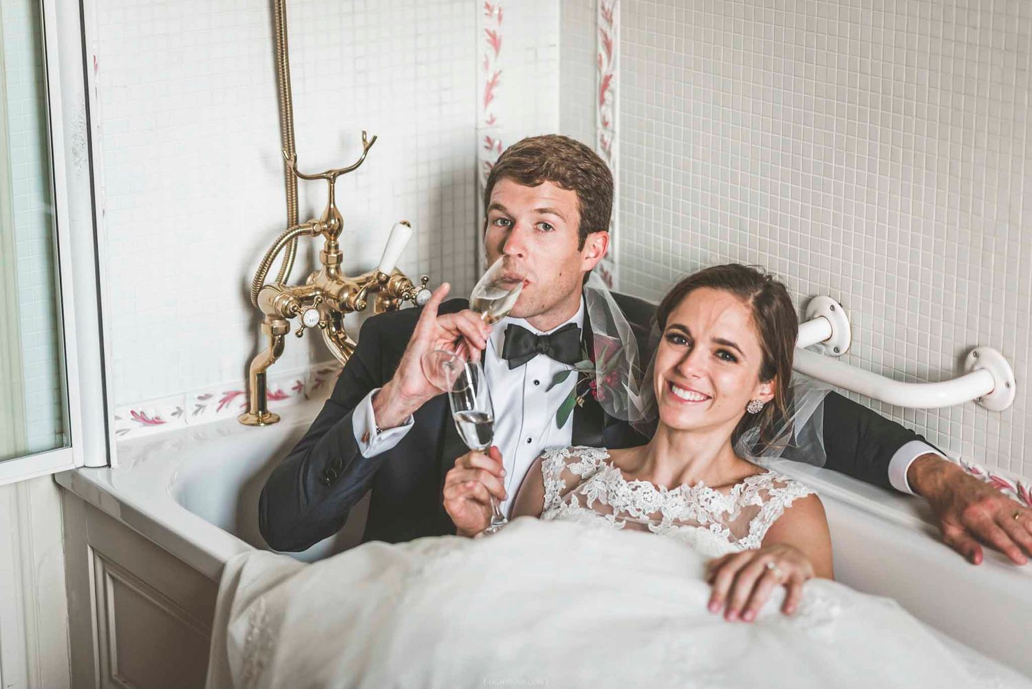 Bride and groom in wedding attire sitting in a vintage bathtub, laughing and holding champagne glasses.
