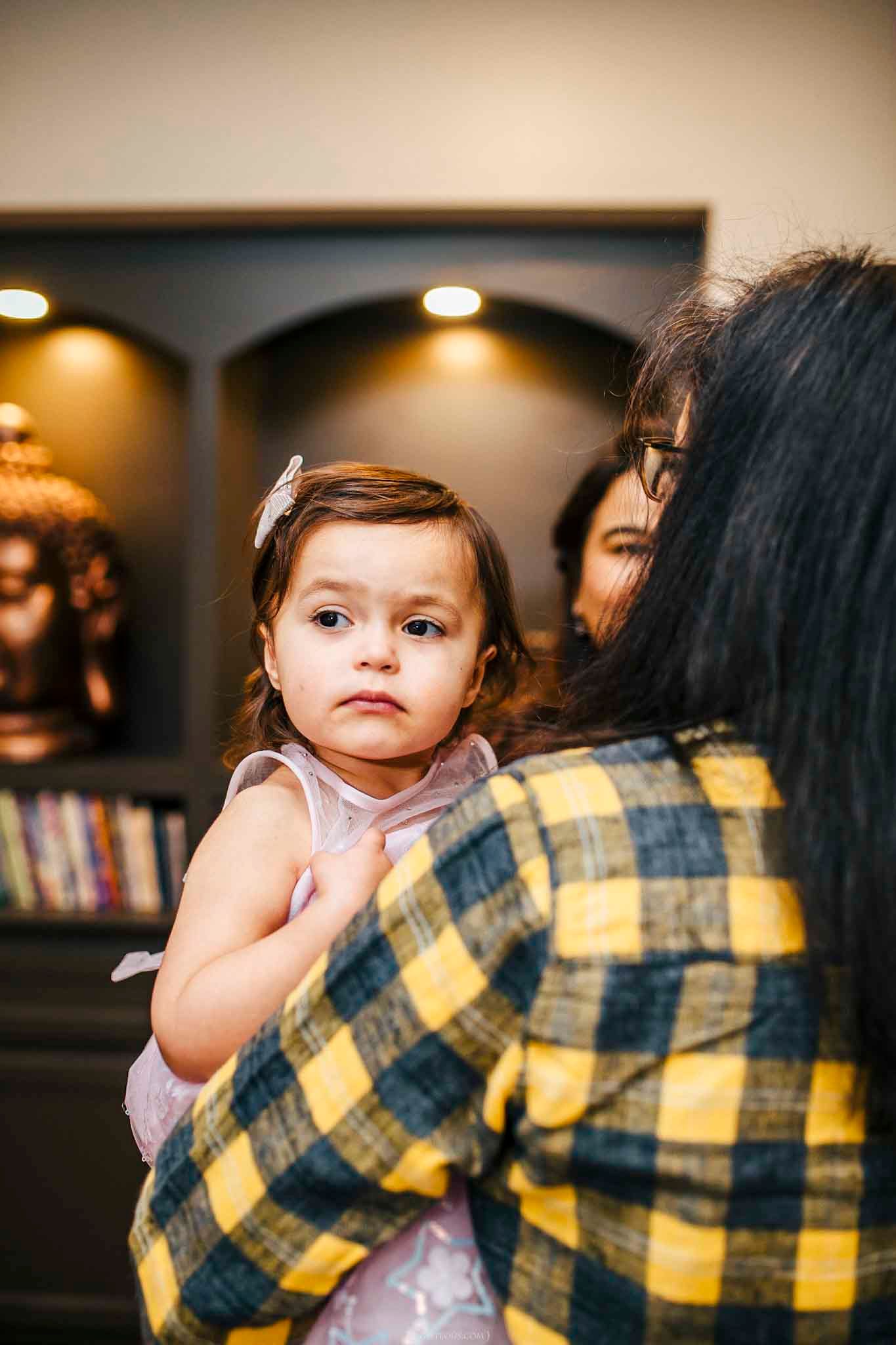 A woman in a yellow plaid shirt holds a cute baby girl in a pink dress inside a warmly lit home.