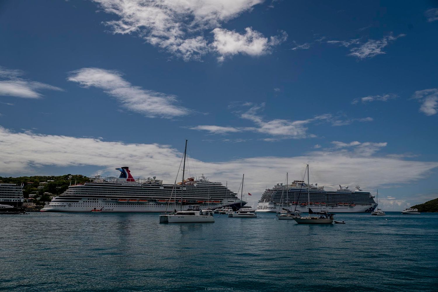 Two large Carnival cruise ships anchored in a tropical harbor surrounded by smaller boats and green hills.