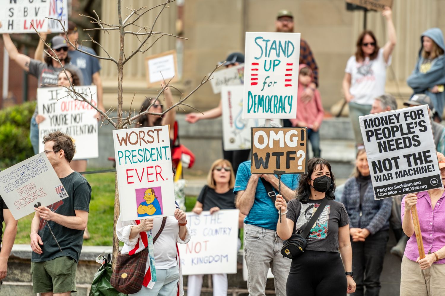 Row of protest signs held up at a political rally with various messages visible.