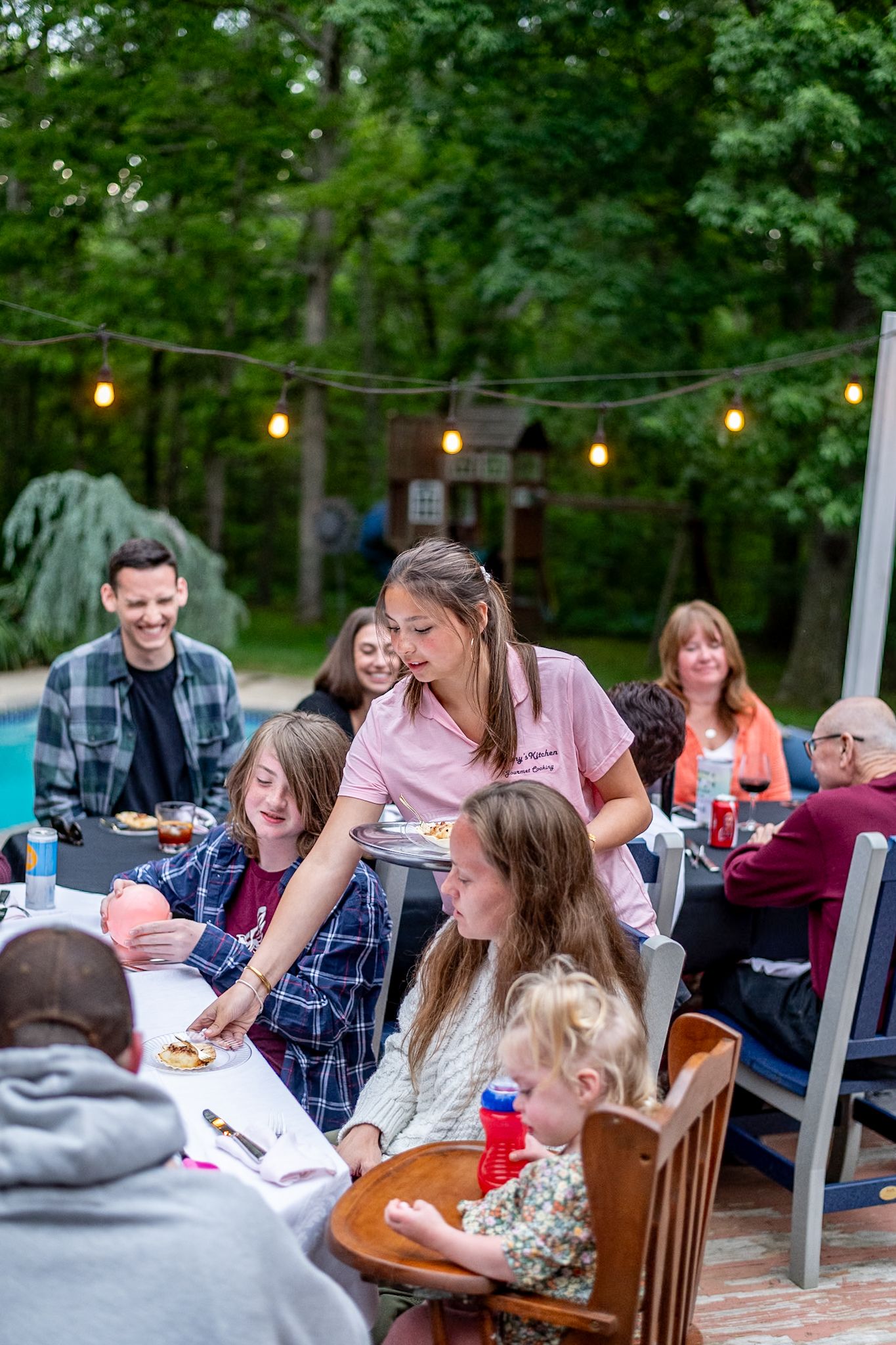 Outdoor gathering with string lights where people sit at tables enjoying food and conversation.
