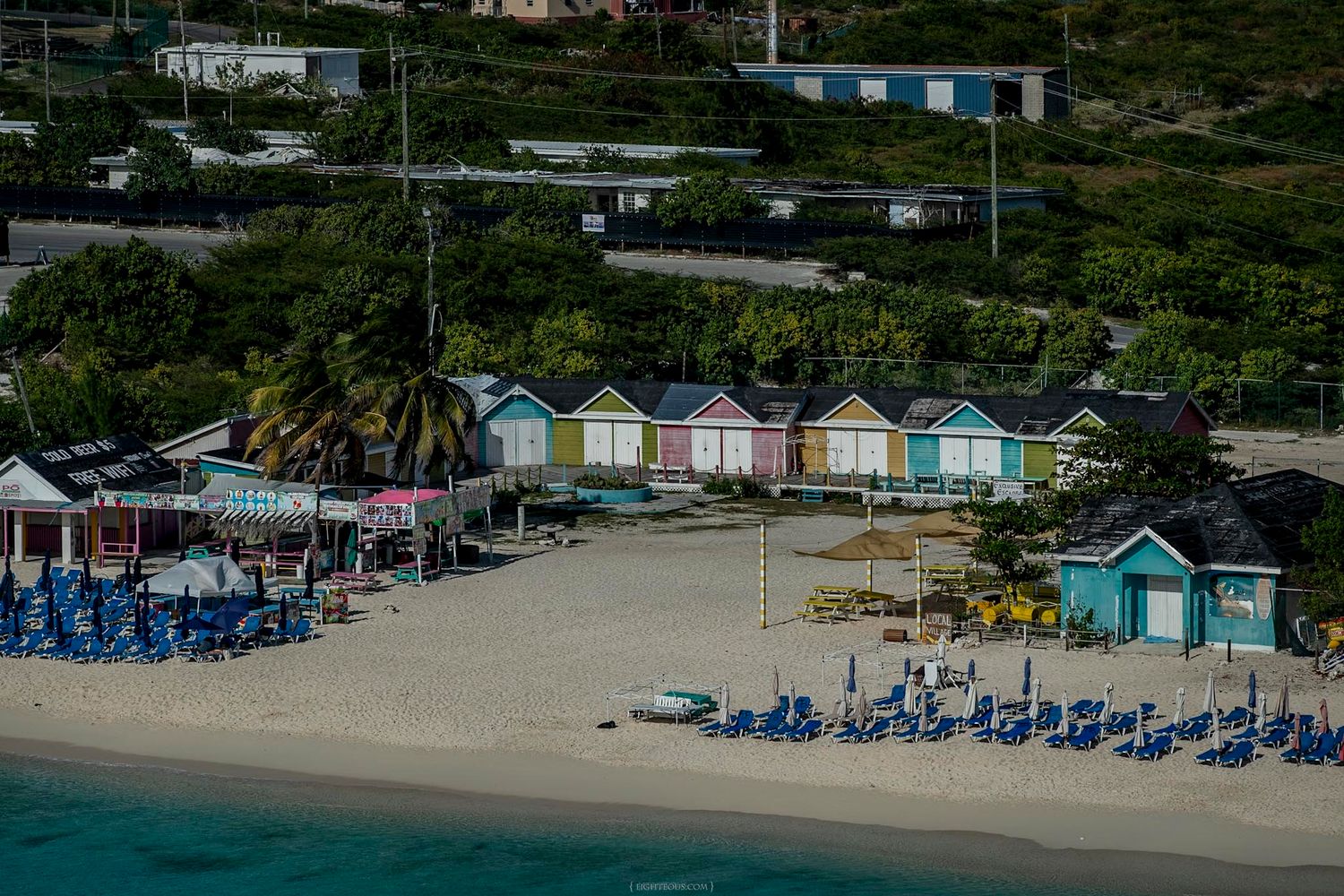 Colorful beach huts line a sandy Caribbean shore with blue lounge chairs and lush green hills in the background.