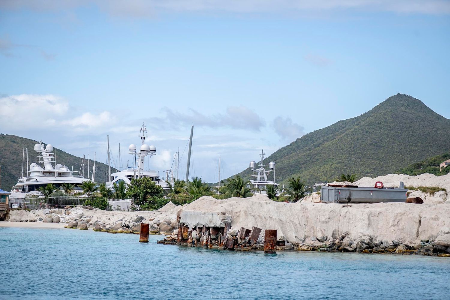 Luxury yachts docked at a marina with rocky breakwater and green mountain backdrop in the Caribbean.