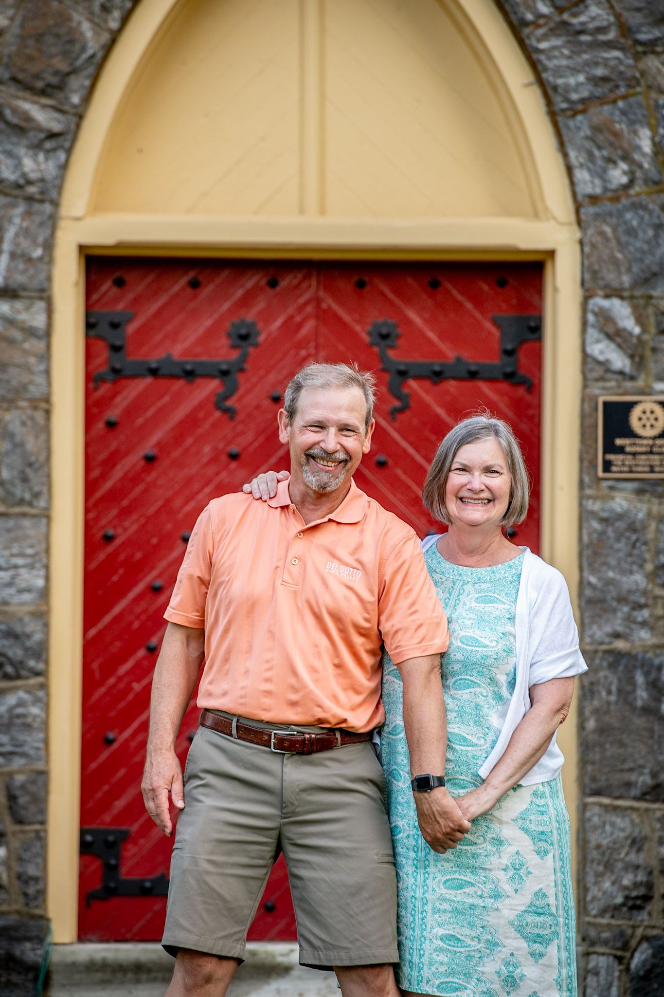 Smiling couple posing in front of a red arched church door, man in orange polo and woman in teal dress.