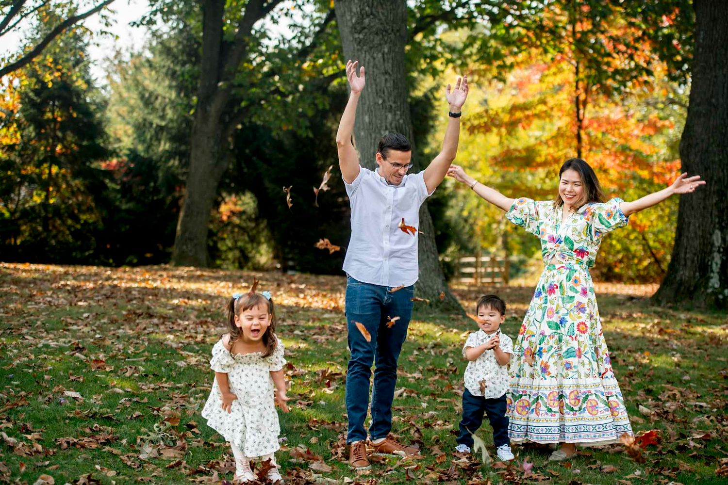 Family of four joyfully tossing autumn leaves in a colorful fall park, parents and two young children laughing outdoors.