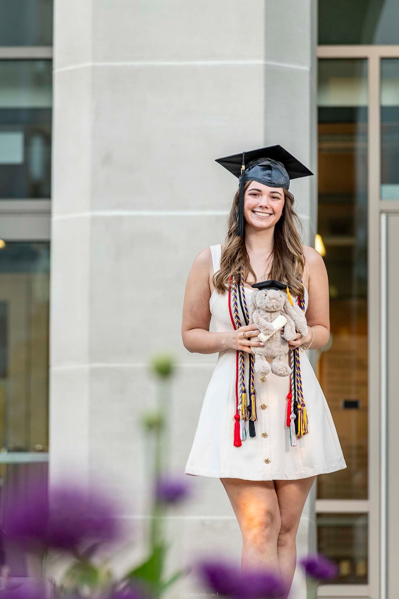 Smiling graduate in white dress and cap holds stuffed animal with colorful honor cords outdoors.