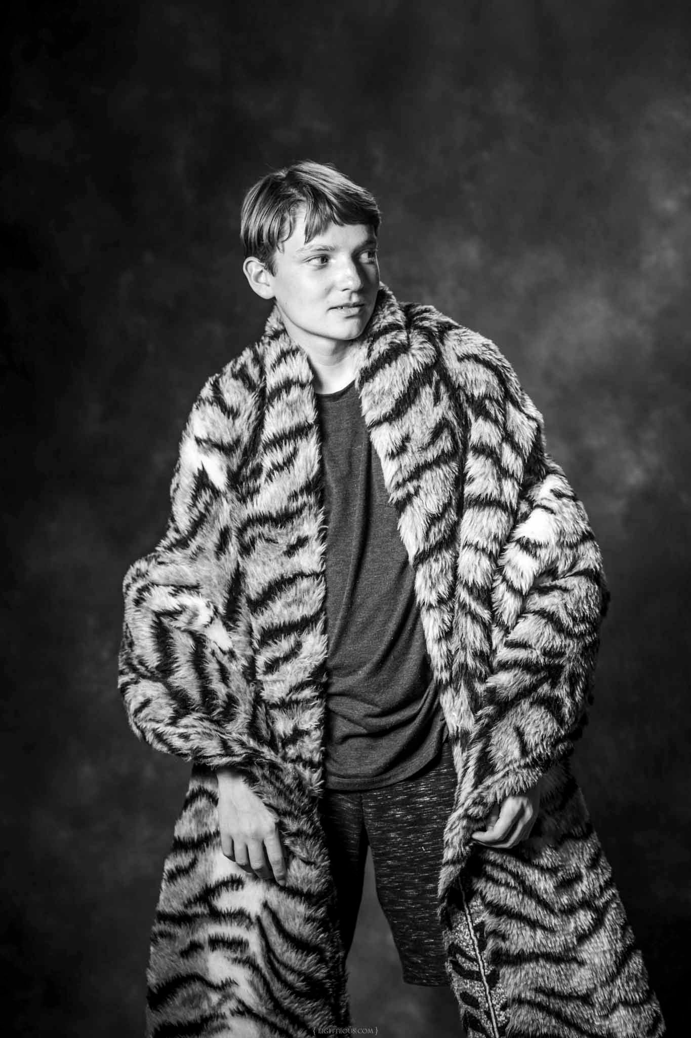 Black and white portrait of a young man wearing an oversized tiger-print fur coat, posing against a dark studio background.
